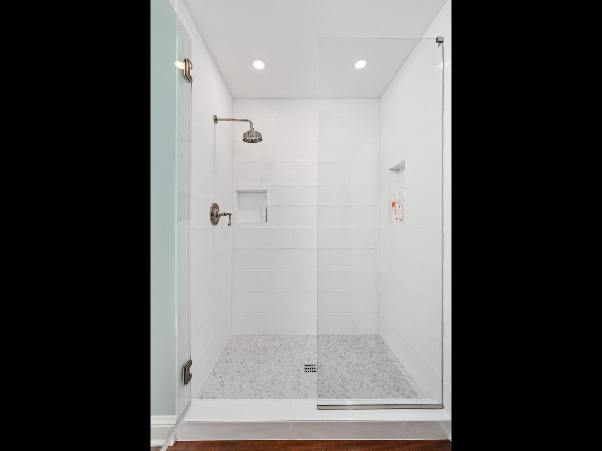 Modern, white tiled shower with a glass door and a rainfall showerhead. The shower has a small built-in niche for toiletries and recessed lighting above. The floor is covered with small, gray tiles.