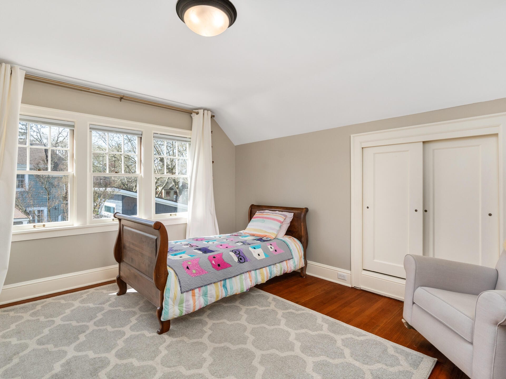 A cozy bedroom with a single wooden bed covered in a colorful quilt featuring animal prints. The room has beige walls, a large window with white curtains, and a light gray armchair. A patterned rug covers the floor. A white closet is in the corner.