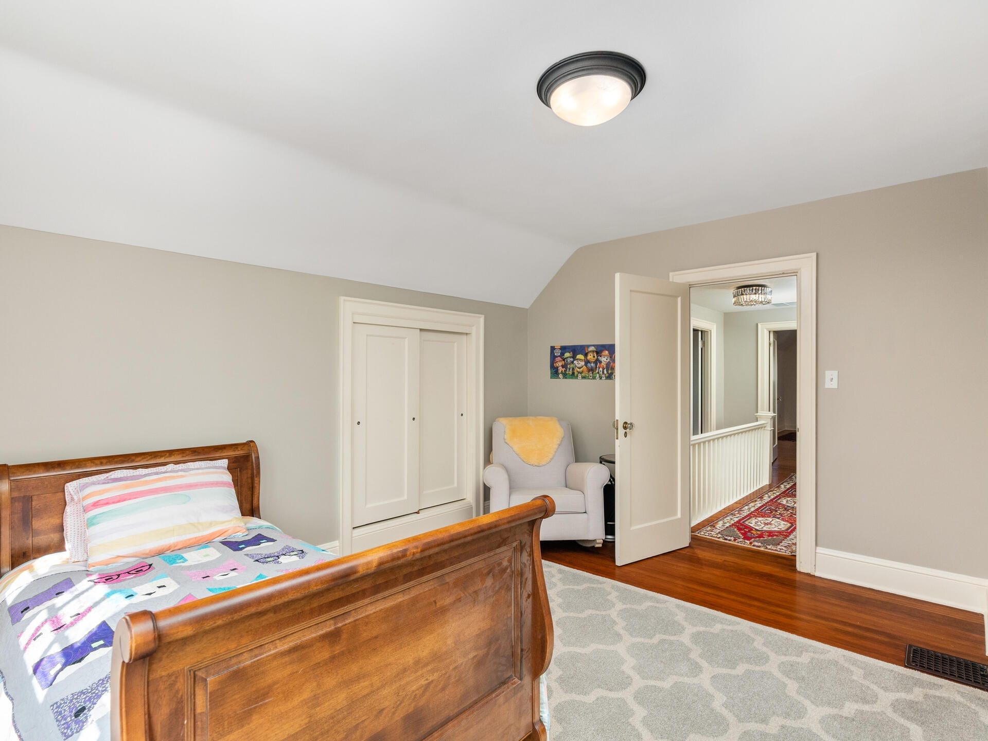 A bedroom with a wooden bed featuring a colorful quilt, a light gray armchair with a yellow blanket, and a gray patterned rug. The walls are light beige, and theres an open door leading to a hallway with a red patterned carpet.