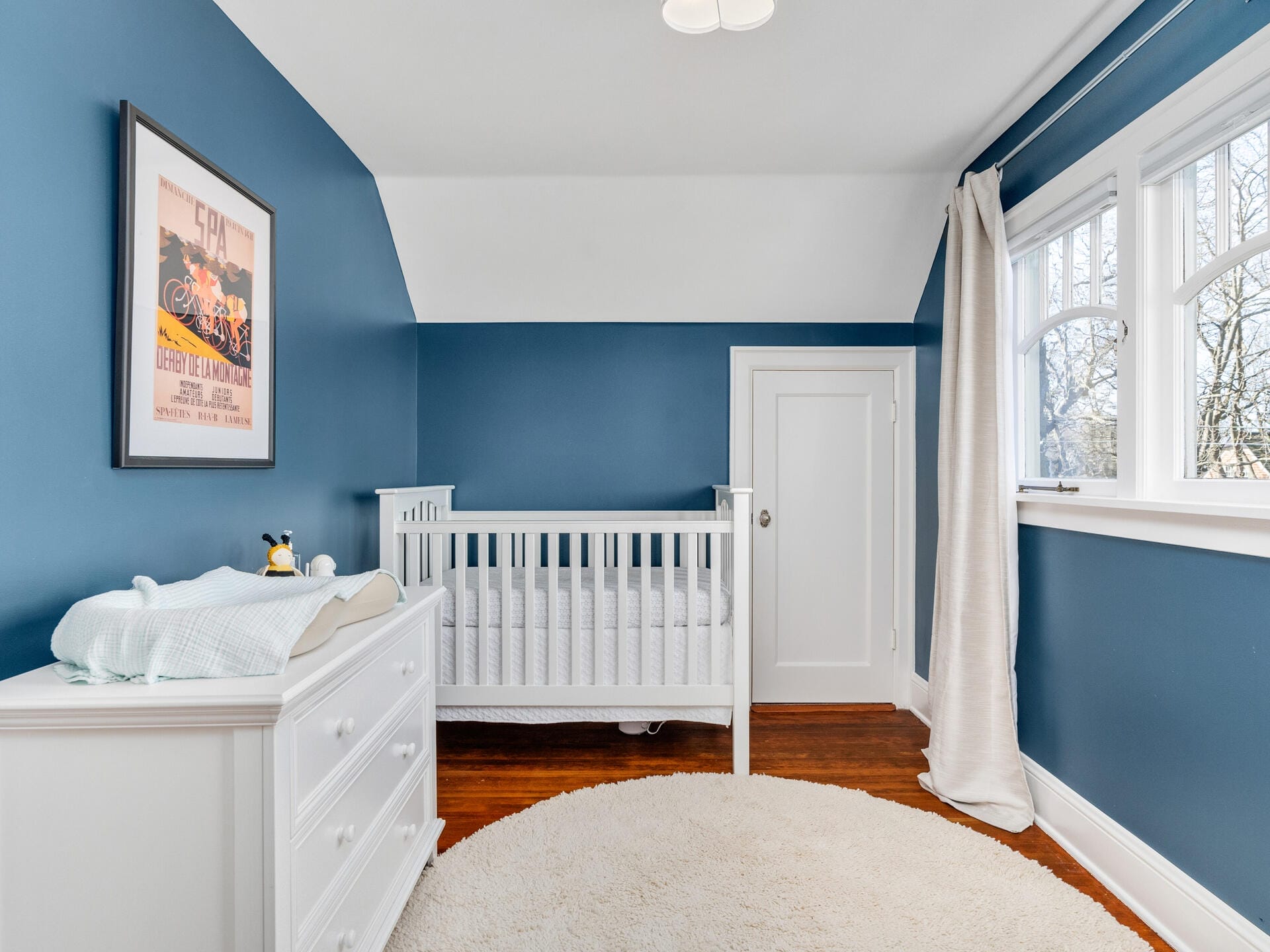 A cozy nursery with blue walls, featuring a white crib and a matching changing table with drawers. A colorful poster is on the wall above the crib. The room has a round beige rug and windows with light curtains, letting in natural light.