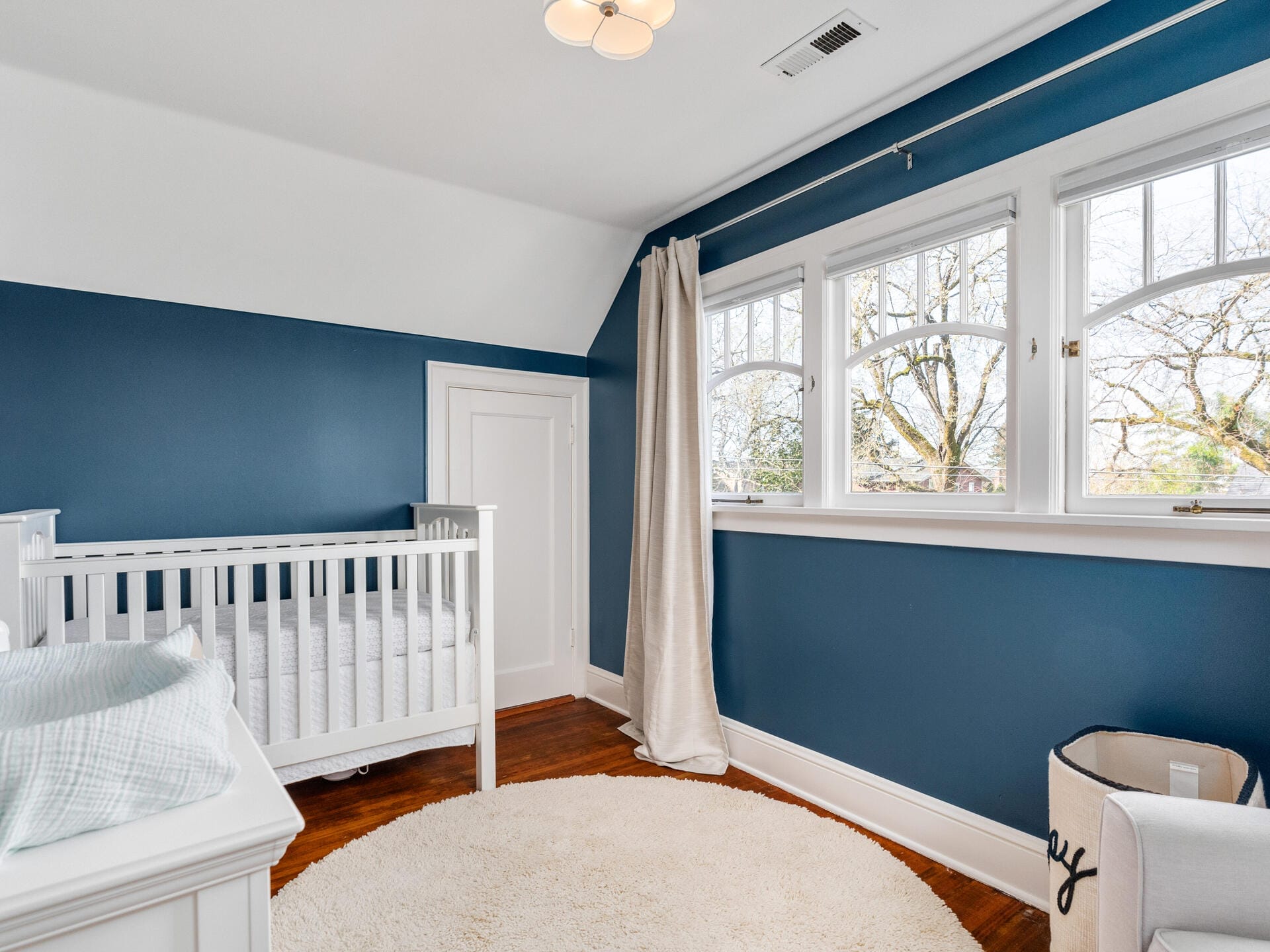 A nursery room with blue walls features a white crib, a changing table, and a round beige rug on wooden flooring. Large windows with white trim allow natural light in, and a cream curtain hangs to the side. A basket sits in the corner.