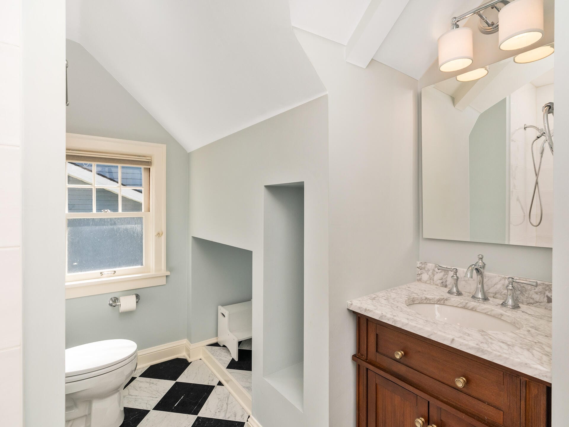 A bathroom with a white toilet, marble countertop sink, and wooden vanity. The floor has black and white tiles. A window is near the toilet, and theres a wall-mounted light fixture above the mirror. The ceiling is slanted.