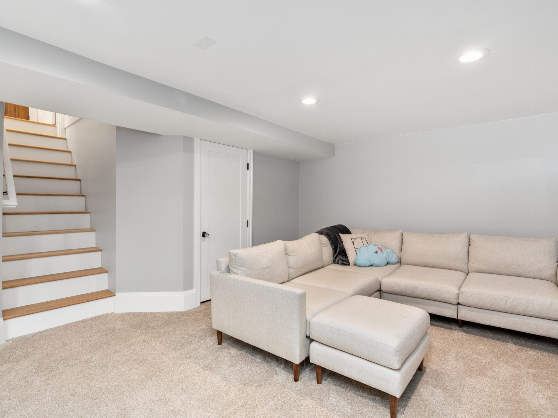 A modern basement with beige carpet features a large L-shaped beige sofa and a light blue pillow. The space is lit by recessed ceiling lights, and a staircase leads upwards on the left. Walls are painted light gray.