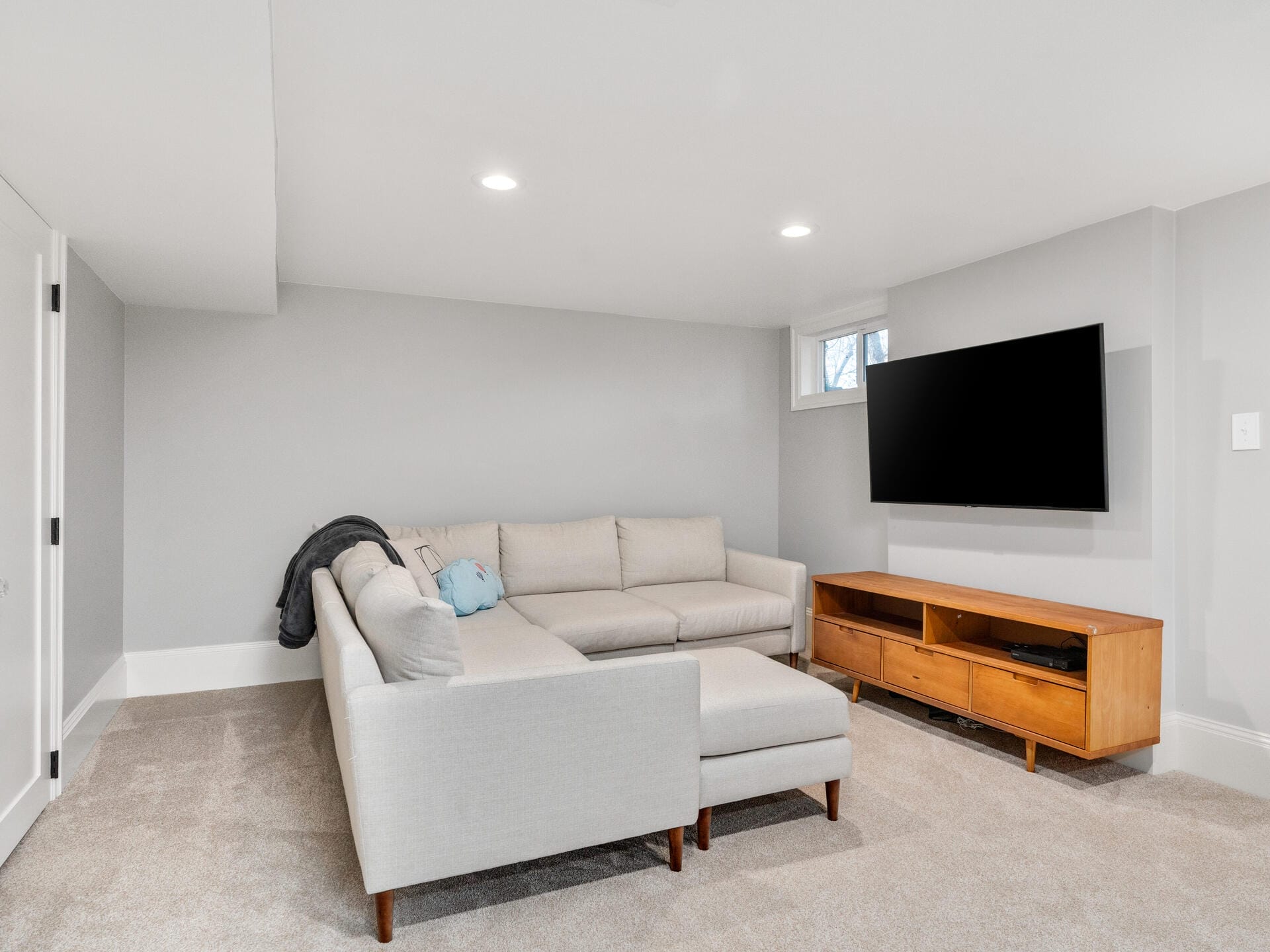 A modern basement living room with light gray walls and carpet. It features a beige sectional sofa with a pillow and a blanket. A flat-screen TV is mounted on the wall above a wooden media console. Small window and recessed lighting provide illumination.