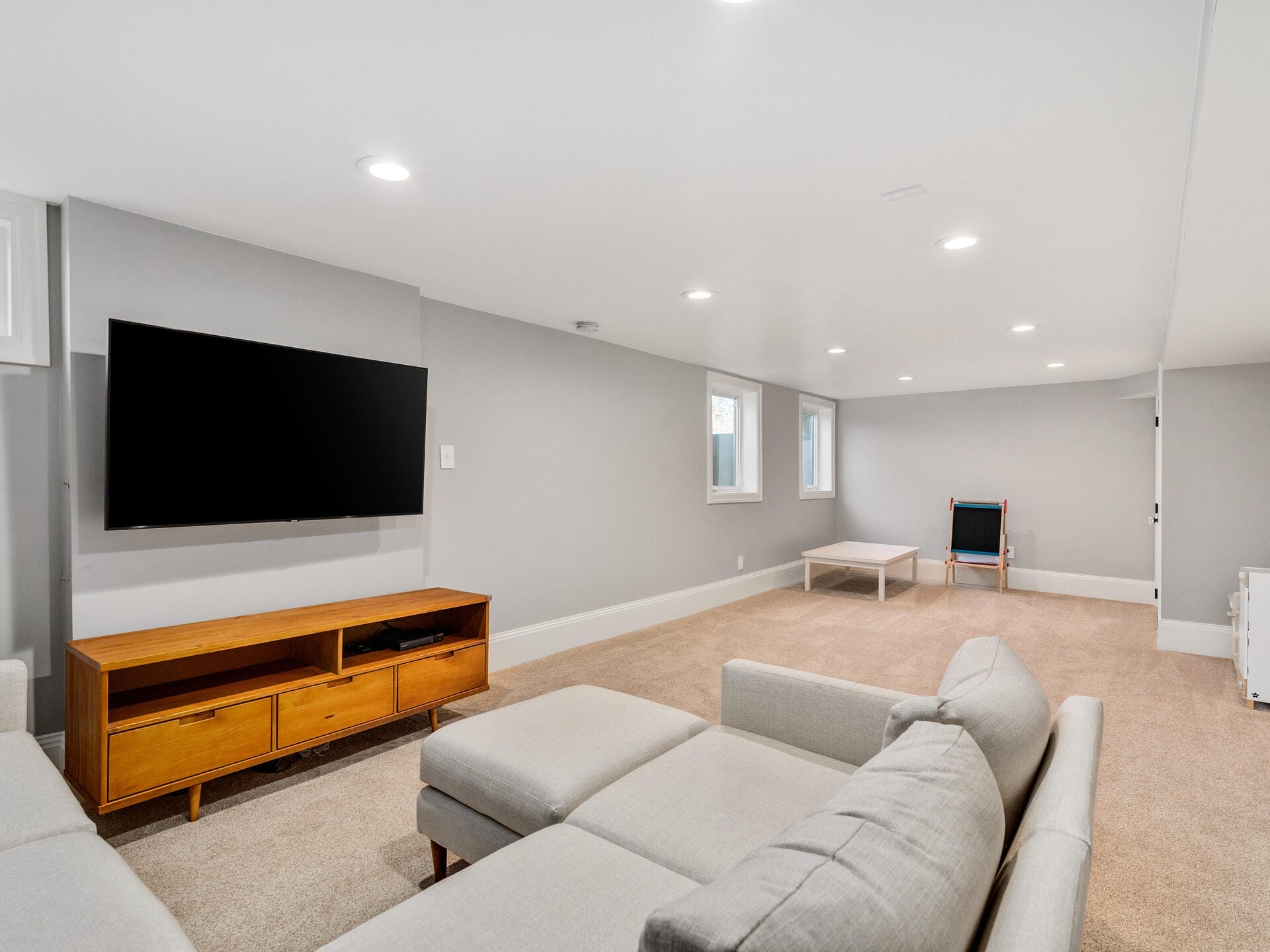A spacious basement room with beige carpet, light gray walls, and recessed ceiling lights. It features a large wall-mounted TV, a light-colored L-shaped sectional sofa, a wooden media console, and a chair with a small table.