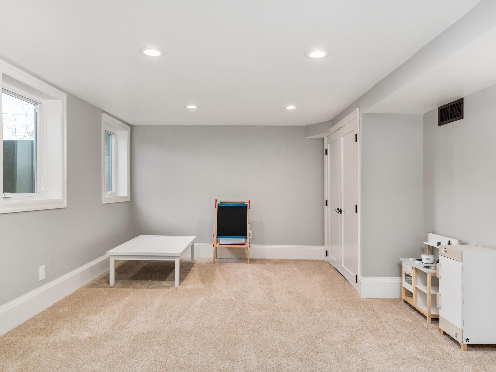 A carpeted room with light gray walls features a small table, a childrens easel, and a play kitchen set. Recessed lighting and two narrow windows provide illumination. A closed double door is on the right side.