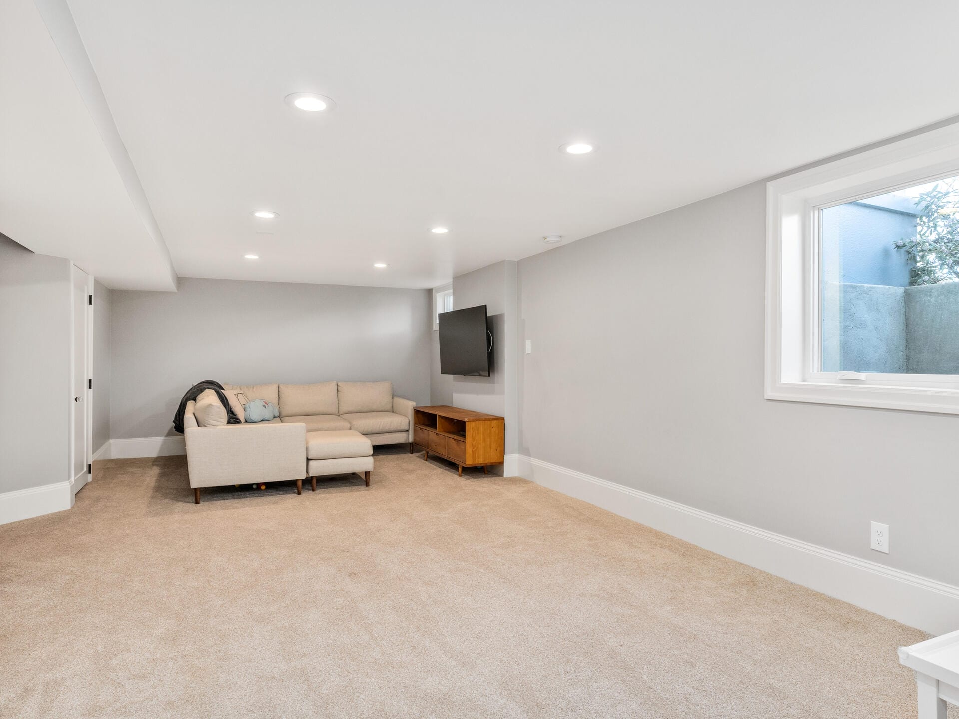 Spacious basement with beige carpet and light gray walls. A sectional sofa and a wooden TV stand are placed along one wall. A large flat-screen TV is mounted on the wall. Natural light enters through a window. Minimalist decor.