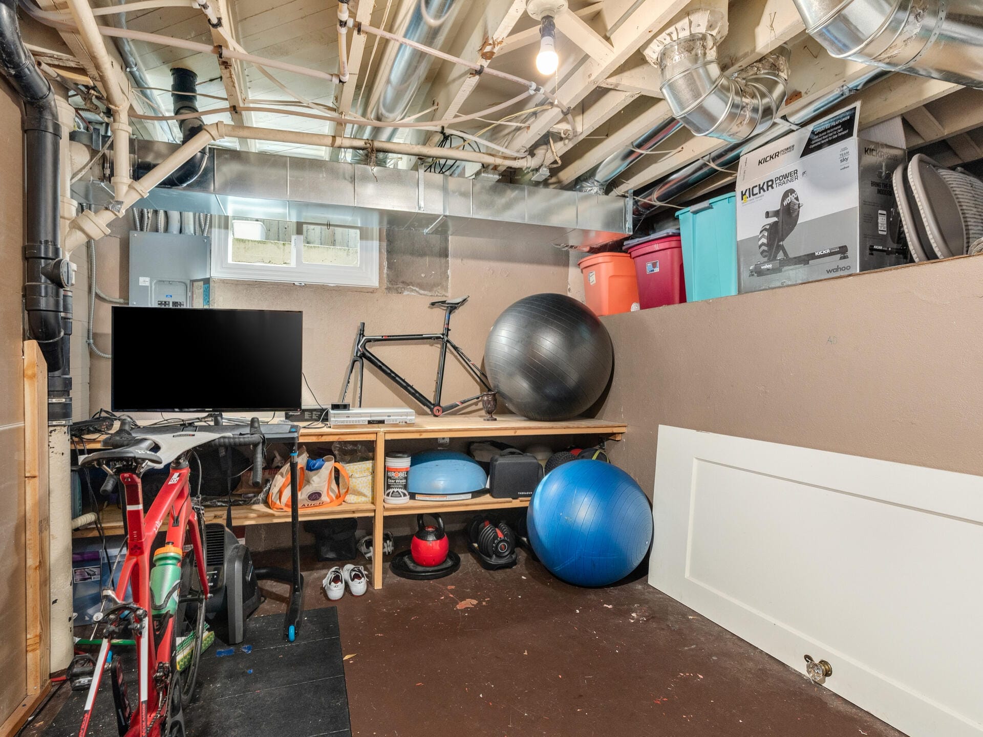 A basement gym with a stationary bike on a trainer, a disassembled bike frame on a shelf, exercise balls, and various equipment stored. Exposed pipes and ductwork are visible on the ceiling.