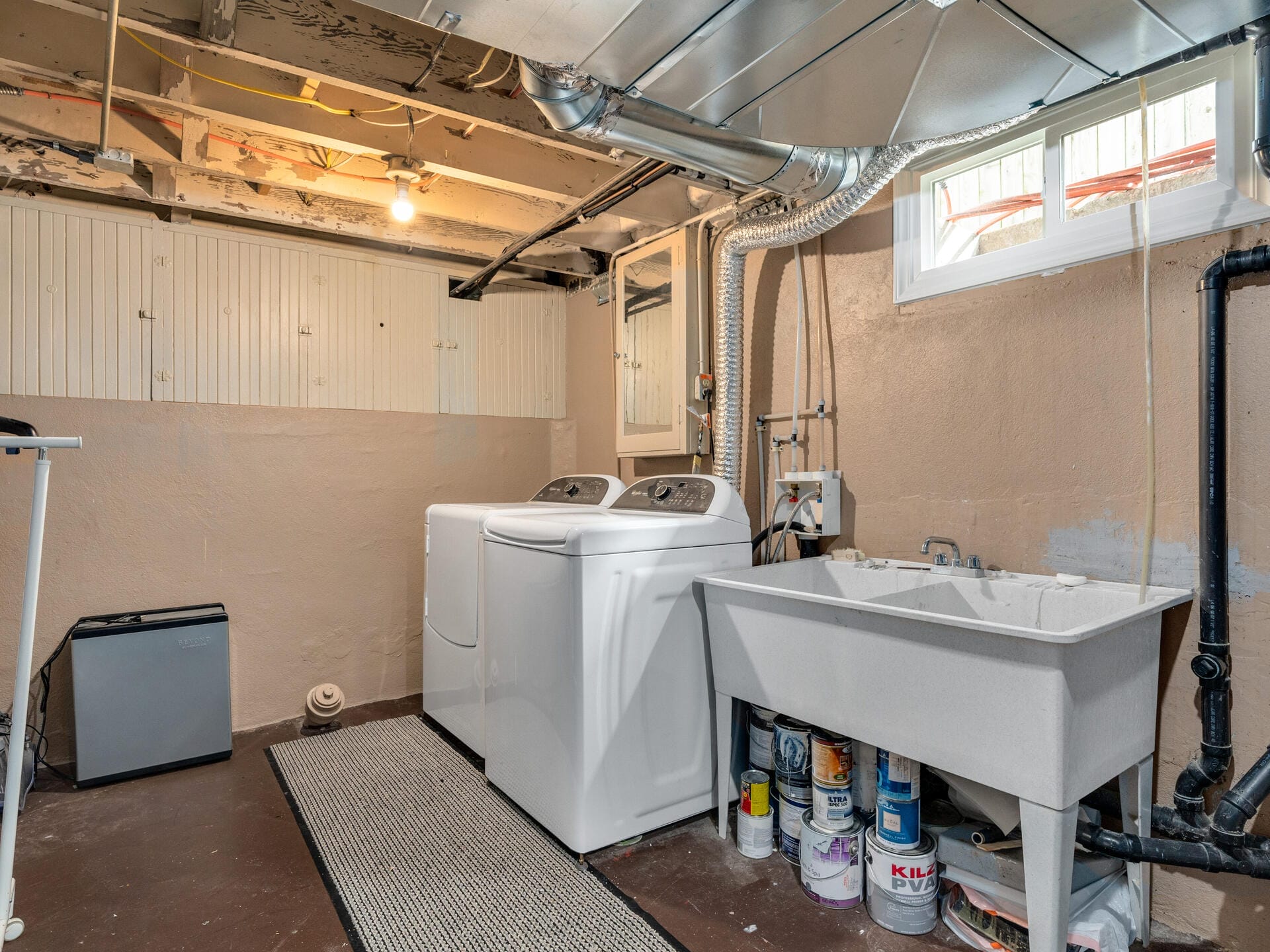 A basement laundry room with exposed beams. Two white washing machines are next to a utility sink. Paint cans are stored underneath the sink. A small window provides natural light. A light bulb hangs from the ceiling, illuminating the space.