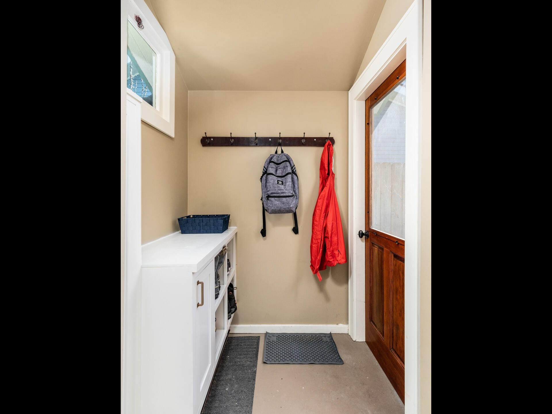 A small mudroom with a wooden door, beige walls, and a white storage bench. A backpack and a red jacket hang from a wall rack above. A gray mat is on the floor. A basket sits on the bench. Natural light filters in through a small window.