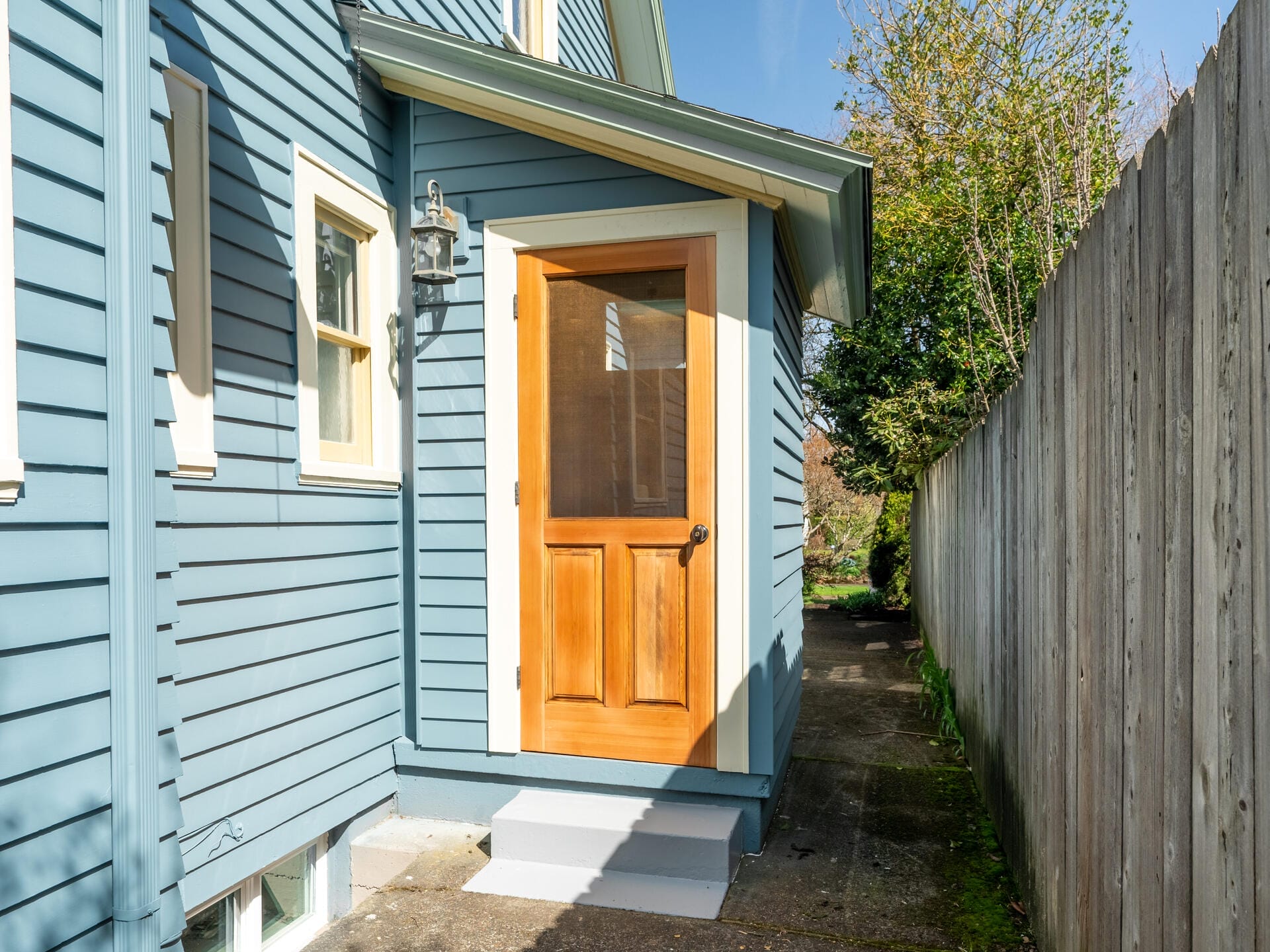 A house with blue wooden siding and a wooden door leading to a small porch. There is a light fixture beside the door and a tall wooden fence running along a narrow path on the right. Lush greenery is visible in the background under a clear sky.