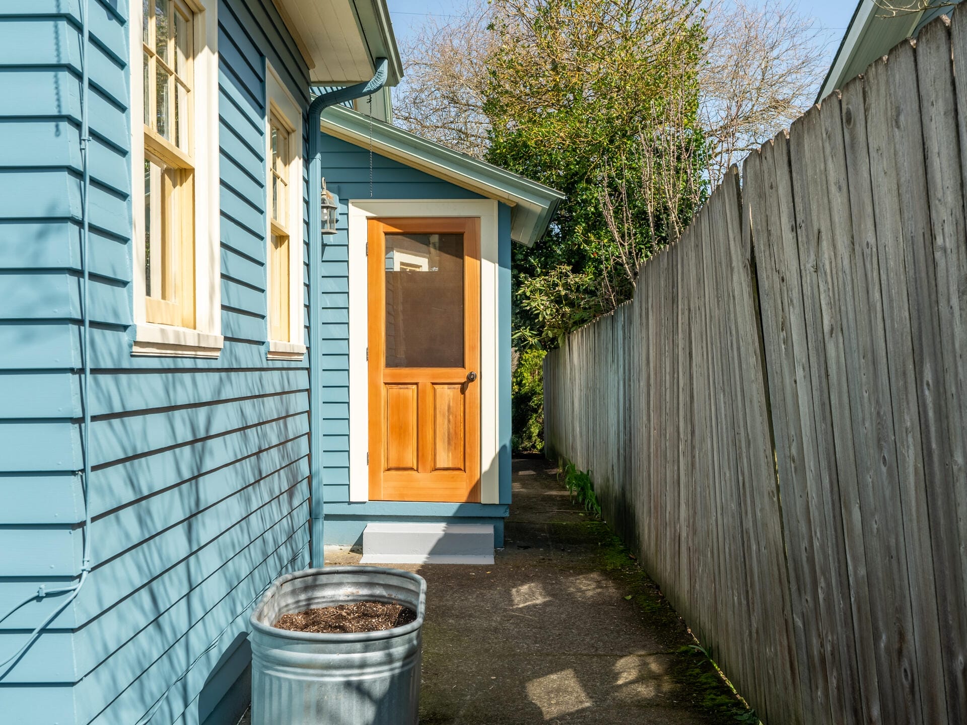 A narrow pathway between a blue siding house and a wooden fence. The house has a wooden door and two windows. A metal planter is placed near the door. Sunlight casts shadows on the pathway and wall. Dense greenery is visible at the paths end.