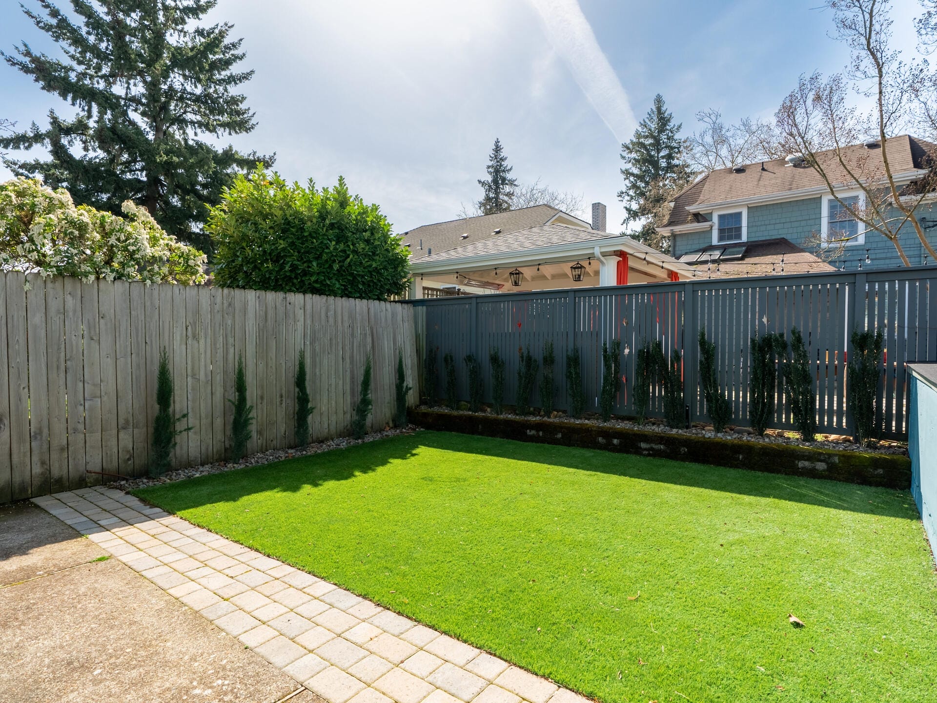 A small backyard with a neatly trimmed lawn and surrounding wooden fence. Several young trees are planted along the fence. Two houses are visible in the background under a clear sky.