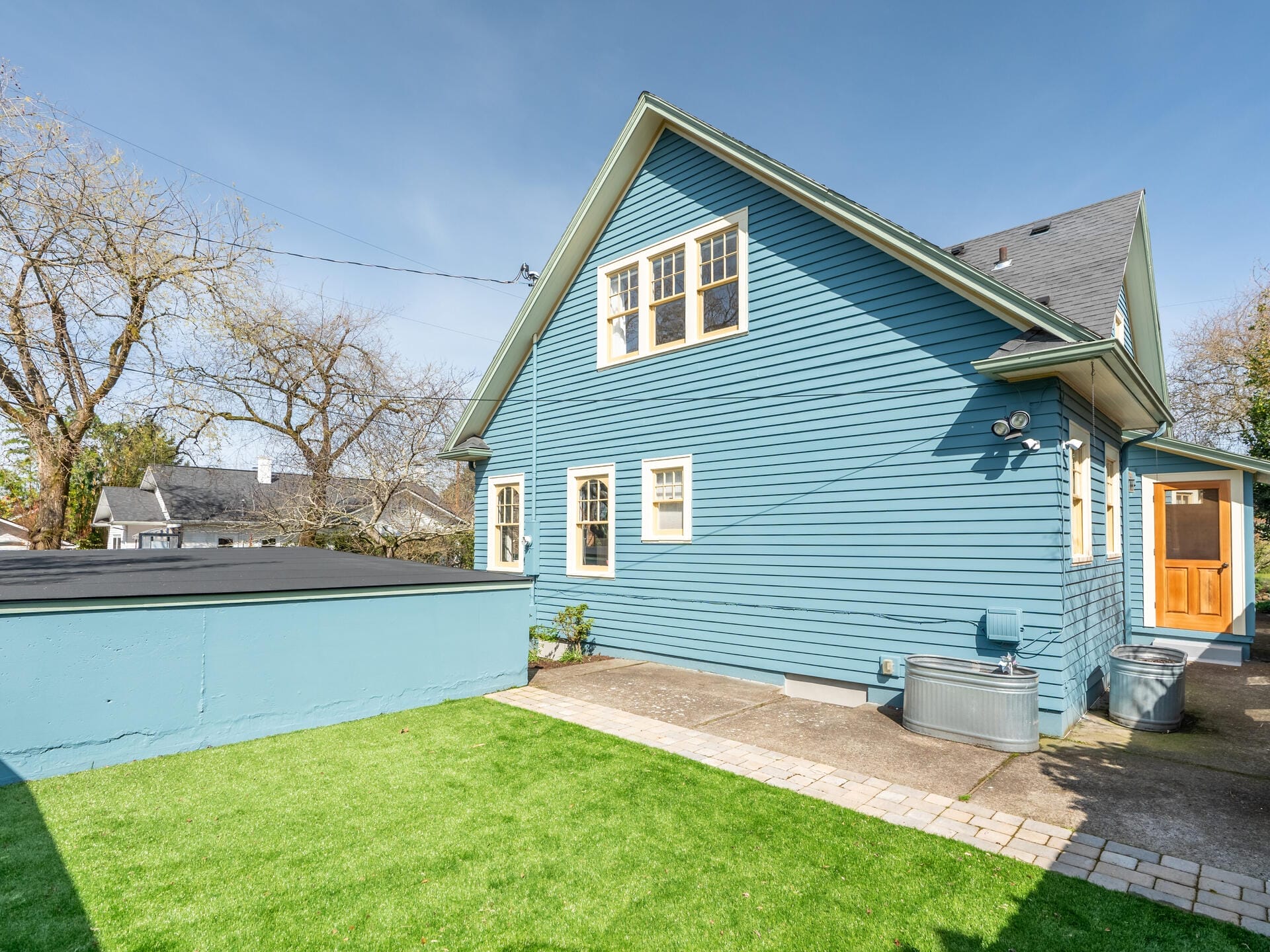 A blue house with multiple windows and a wooden door, surrounded by a small concrete patio and a well-manicured lawn. The sky is clear, and some bare trees are visible in the background.