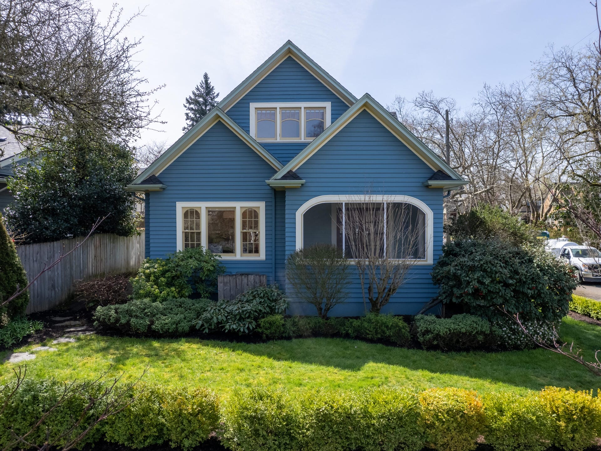 A charming blue house with white trim, featuring a gable roof and a front porch. Surrounded by a well-maintained garden with green shrubs and trees, and a wooden fence on the left. A clear sunny day with a serene neighborhood setting.