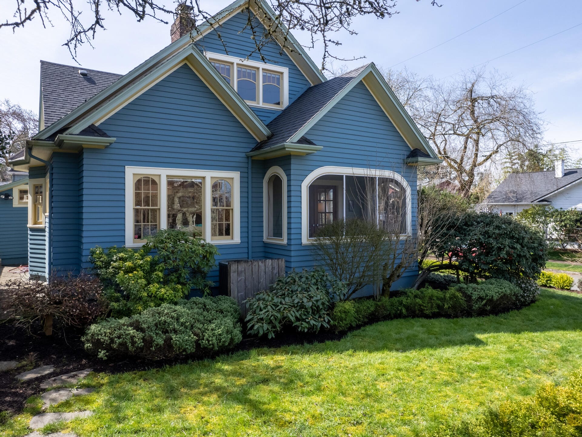 A charming blue house with a gable roof is surrounded by lush greenery and shrubs. The windows are partially shaded by trees, and a well-manicured lawn extends in front, reflecting a sunny day.