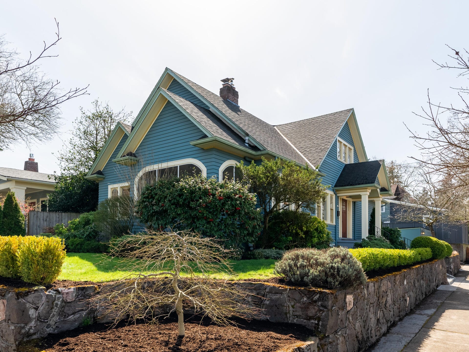 A charming, blue craftsman-style house with a sloped roof and white trim sits behind a well-kept garden. The garden features bushes, a small tree, and stone retaining walls, all under a clear sky.