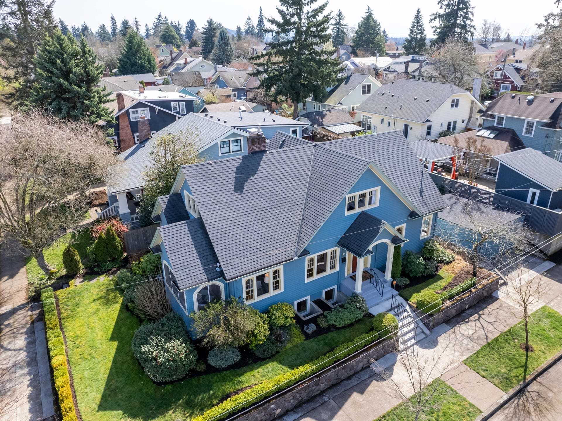 Aerial view of a blue house with a dark roof surrounded by a neatly landscaped yard. The neighborhood features similar houses, trees, and well-maintained greenery. The sky is clear, suggesting a sunny day.