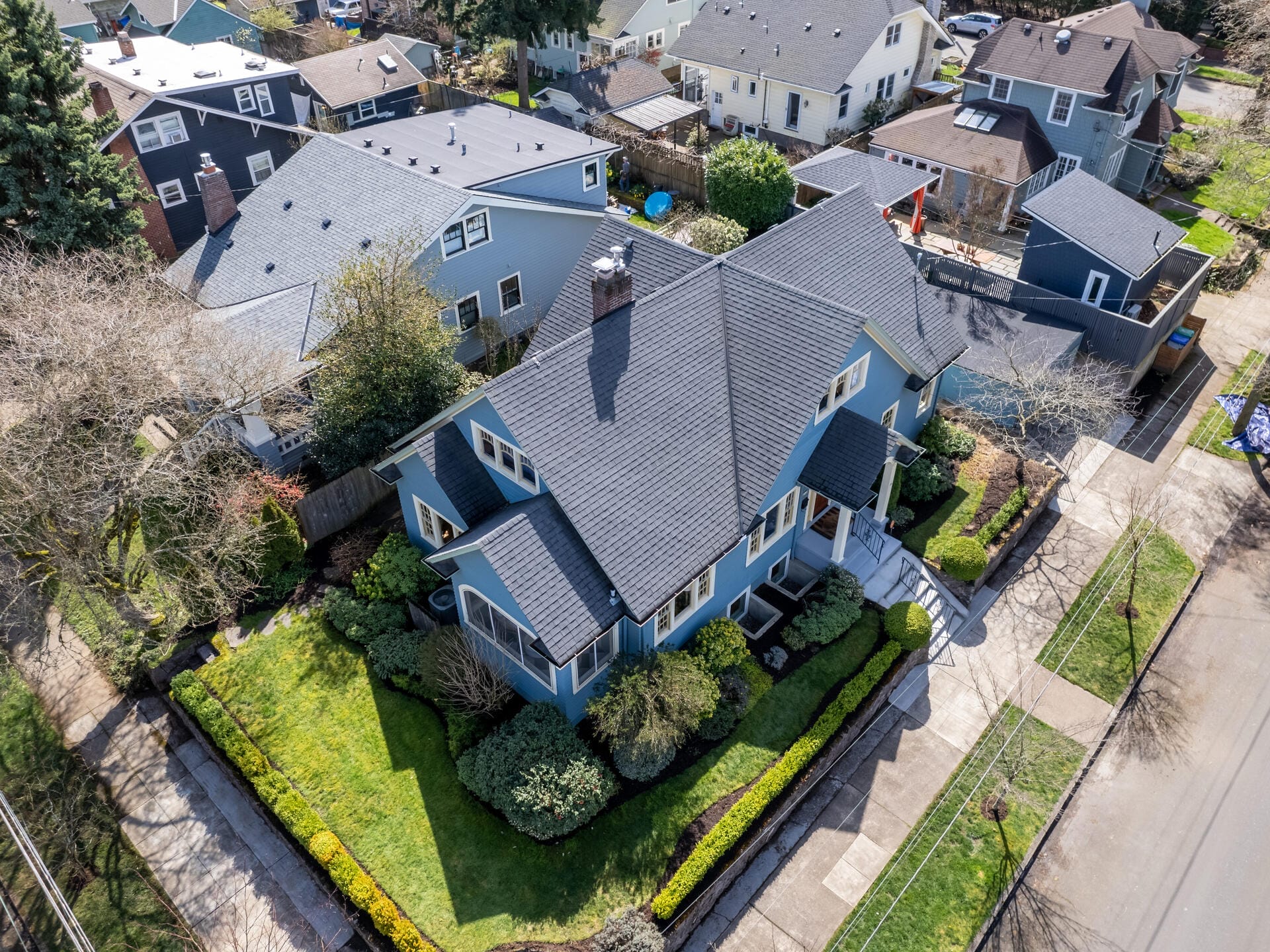Aerial view of a charming blue house with a dark gray roof, surrounded by a neatly manicured lawn and hedges. Nearby are other similar homes, tree-lined streets, and a few mature trees. The scene is residential and peaceful.