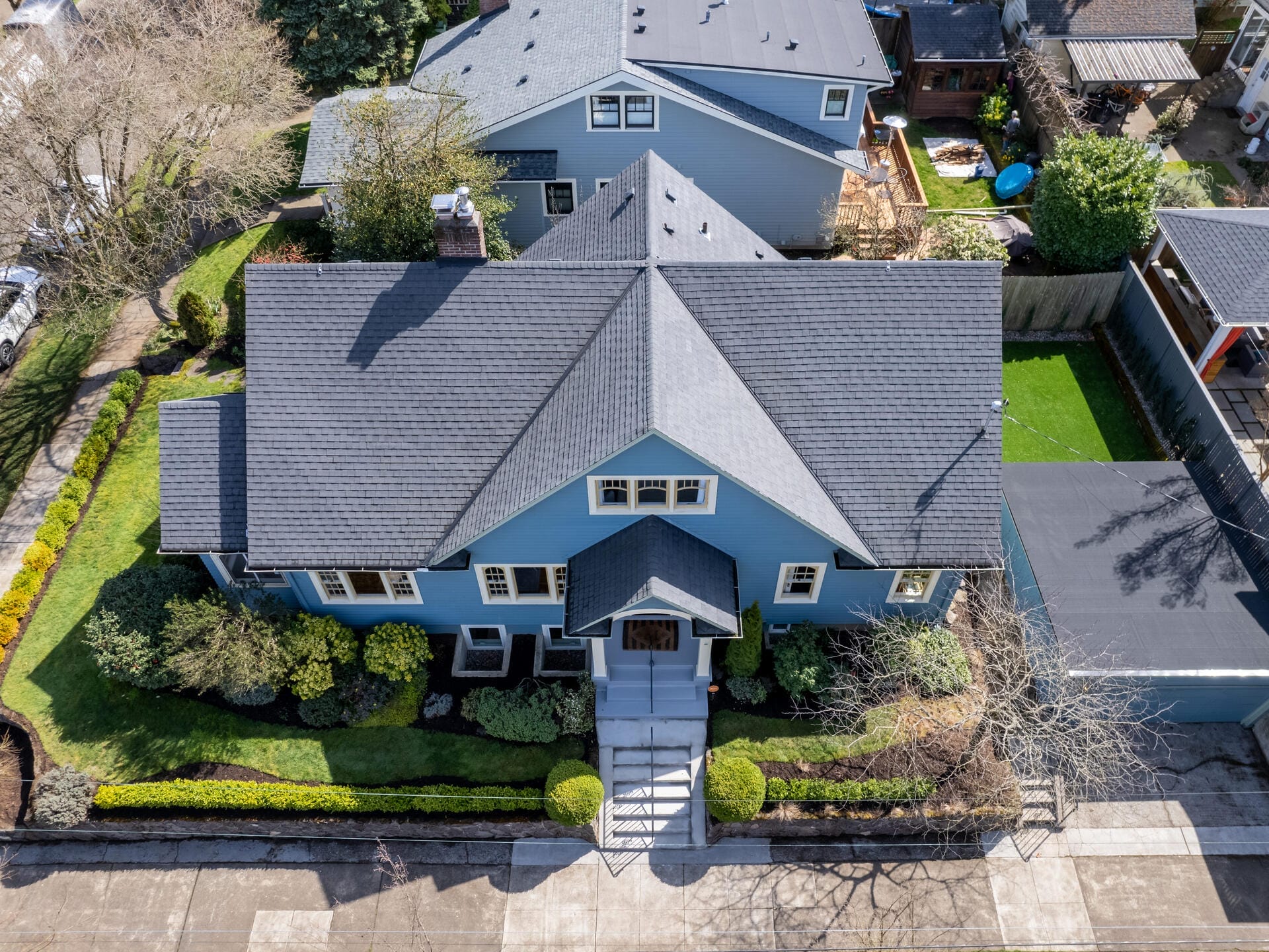 Aerial view of a blue house with a dark gray roof, surrounded by a green lawn and neatly trimmed bushes. The property includes a driveway and is situated on a tree-lined street.