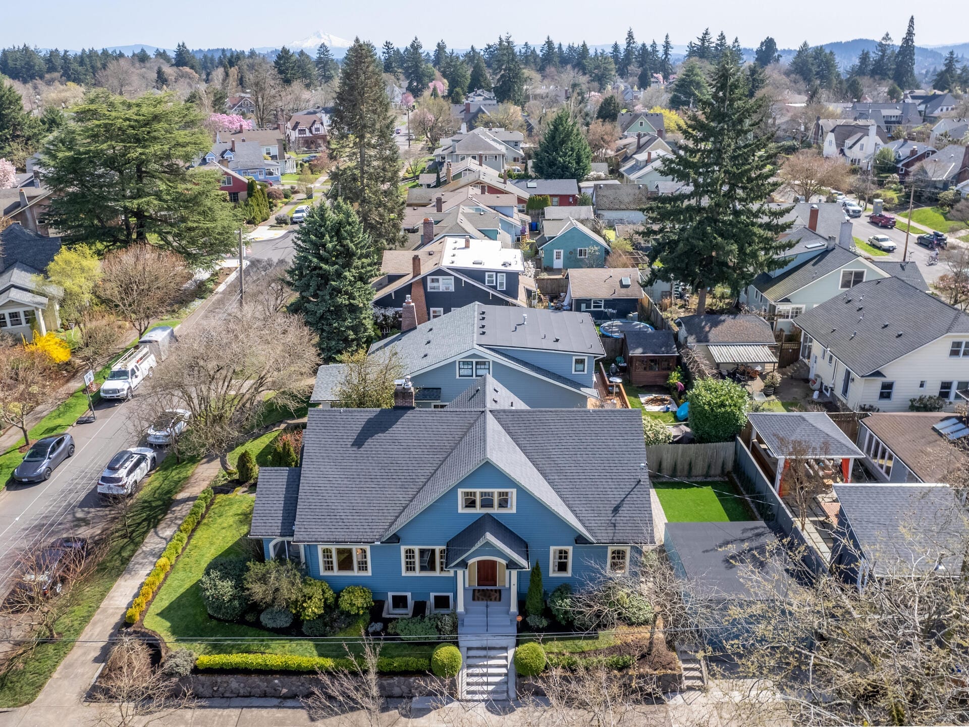 Aerial view of a suburban neighborhood featuring a row of houses on a tree-lined street. The foreground shows a large, blue two-story home with a neatly maintained yard and driveway. Surrounding homes are diverse in style, with lush greenery throughout.