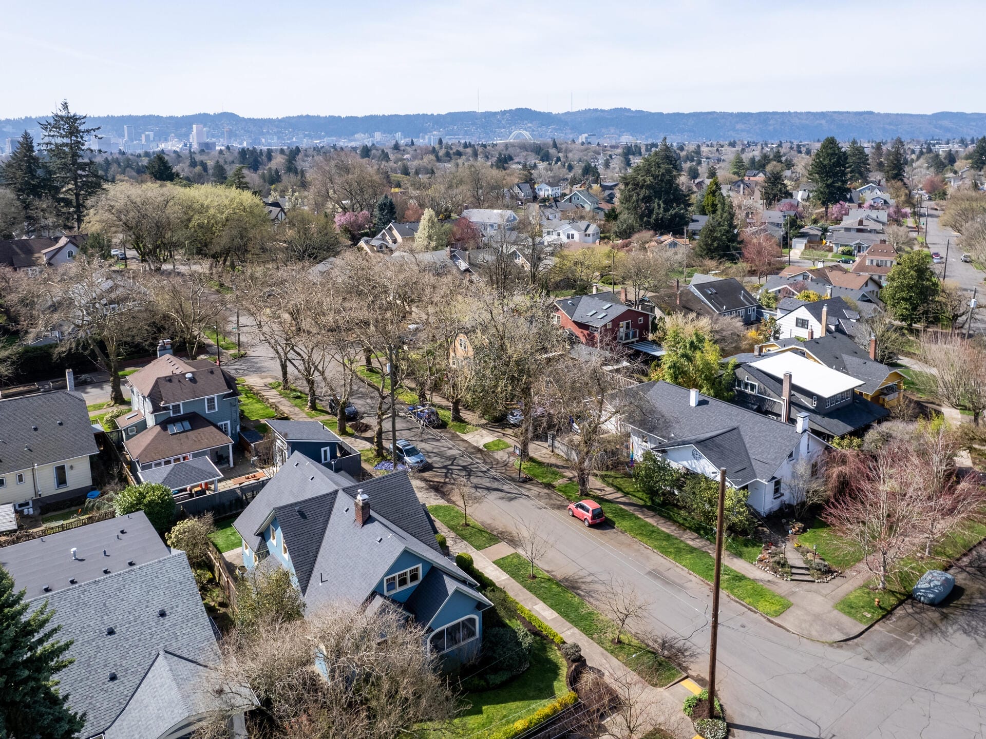 Aerial view of a suburban neighborhood with tree-lined streets and houses in various styles. The landscape is dotted with greenery and patches of blossoming trees, set against a backdrop of distant hills under a clear sky.