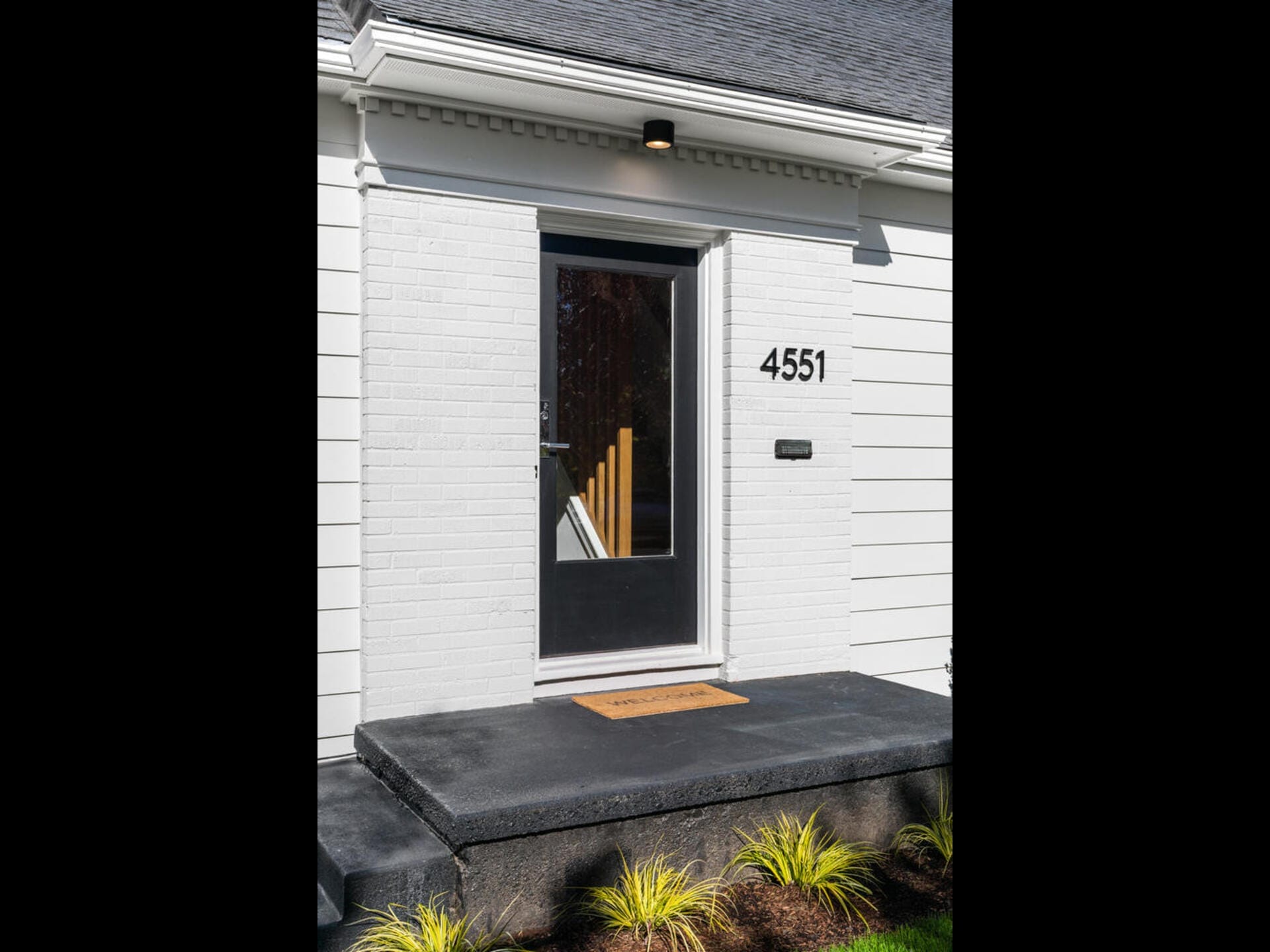 A modern white house entrance with a black steel door featuring glass panes. The house number, 4551, is displayed on the wall. A simple doormat lies on a dark step, and small ornamental plants are at the base.