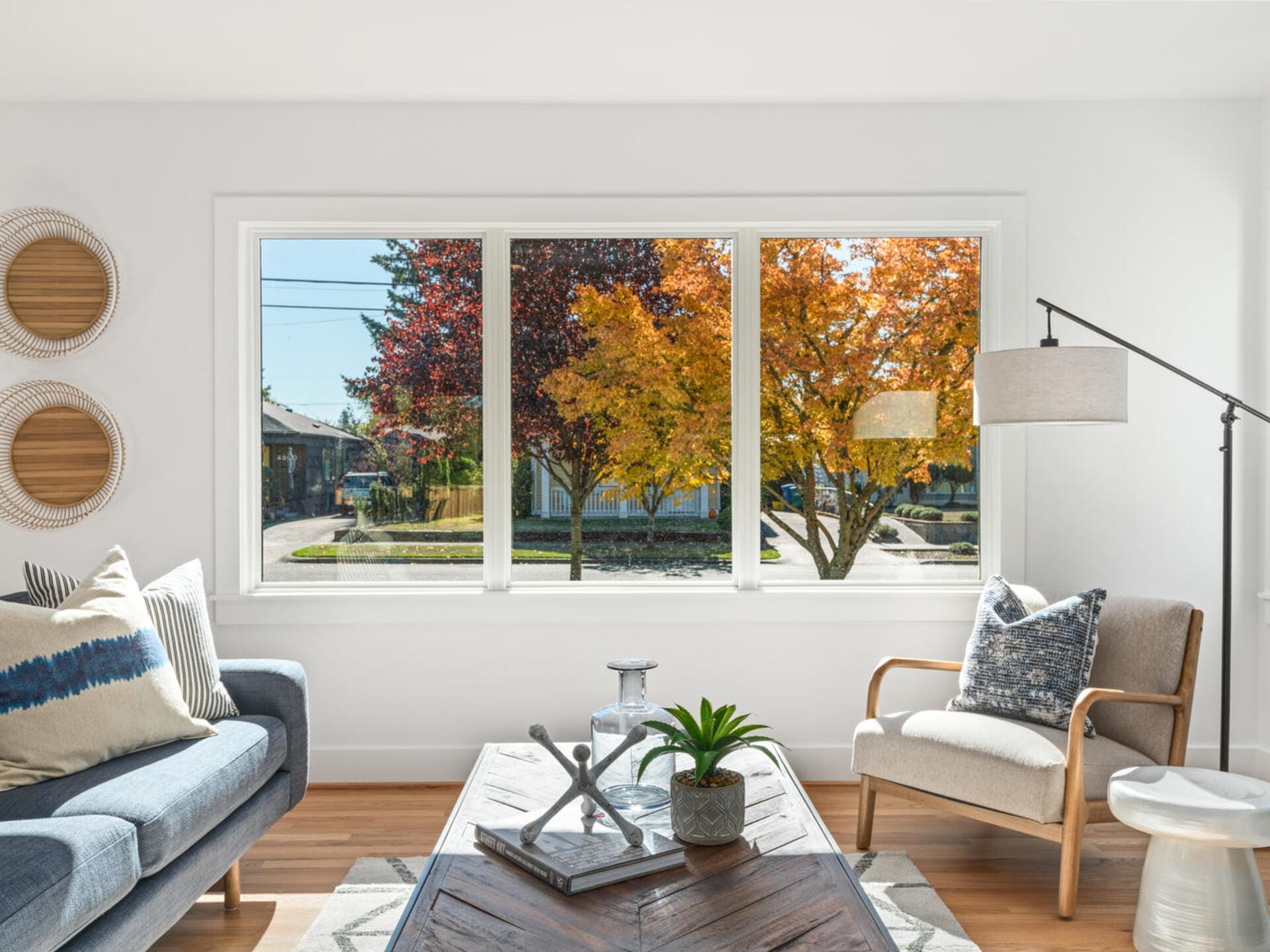 A bright living room with a large window showing colorful autumn trees outside. The room features a gray sofa, an armchair, and a wooden coffee table with a plant and books. A tall lamp stands nearby, and woven wall decor is visible.