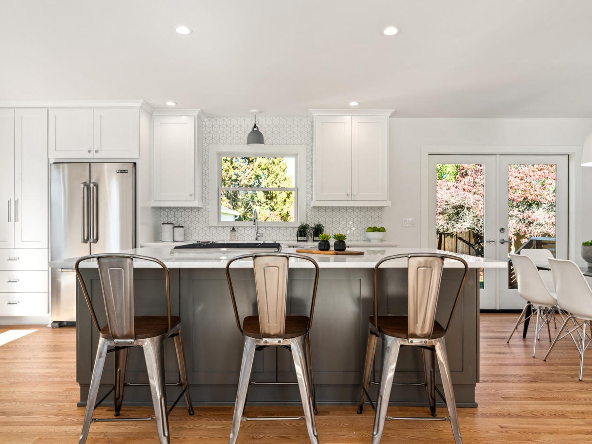 A modern kitchen with a center island, featuring bar stools, white cabinets, and stainless steel appliances. The room is bright, with large windows and glass doors leading outside. A small dining area is in the background.