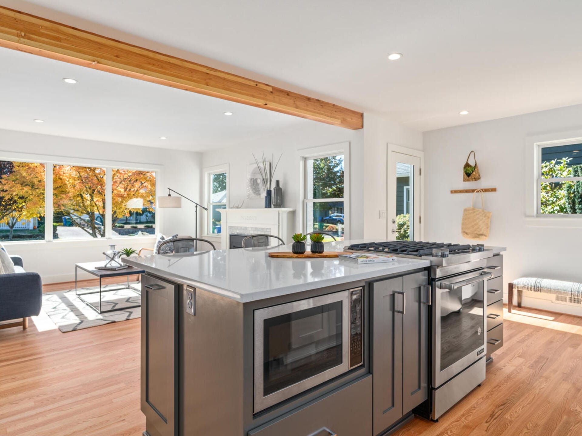 Modern kitchen and living area with hardwood floors. The kitchen features a large island with a stove and microwave. The living room has a gray sofa and a wood beam across the ceiling. Large windows reveal trees with autumn leaves outside.