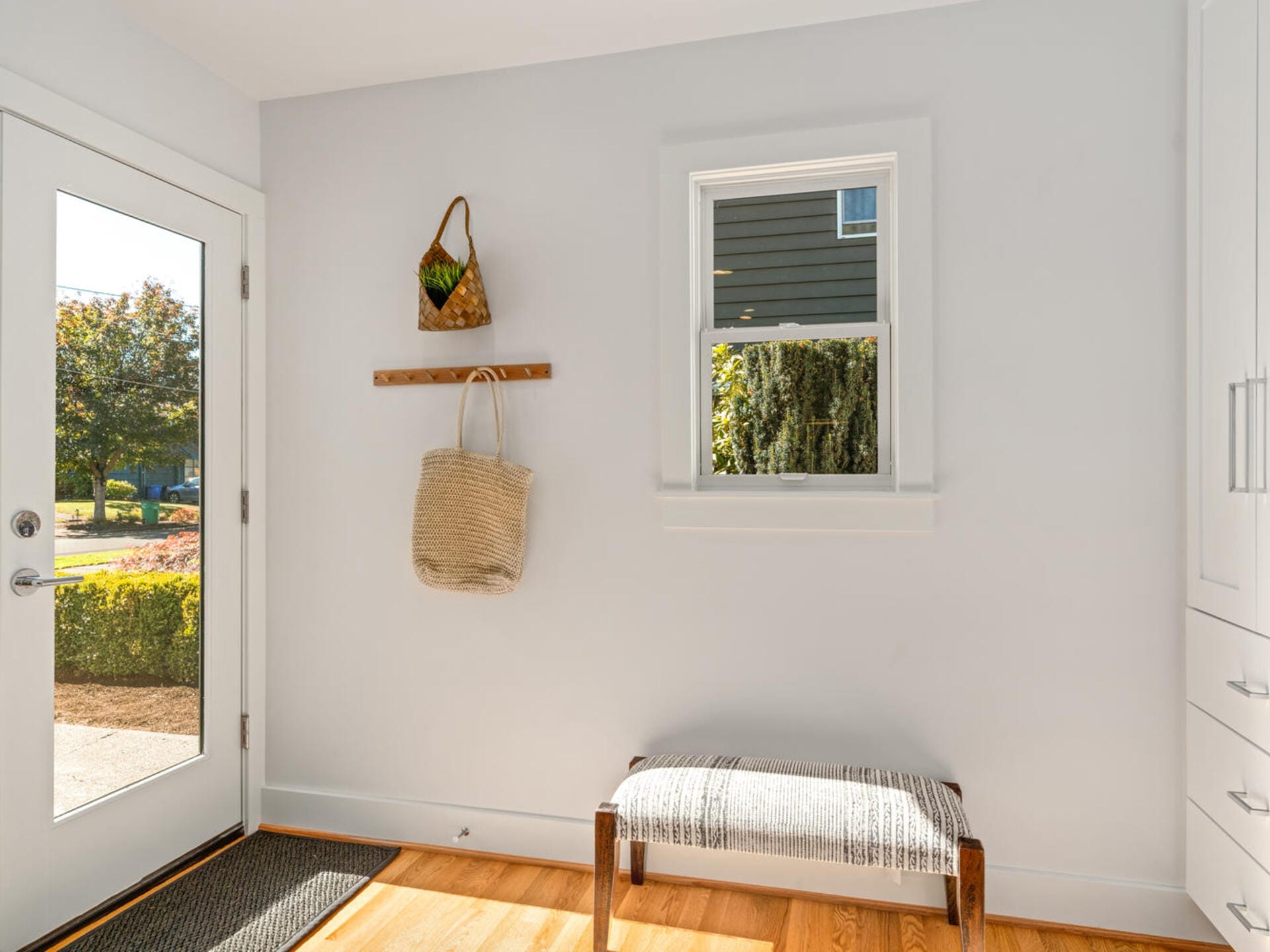 A bright entryway with a glass door on the left, a window on the right, and a striped bench below. A woven basket and a wicker bag hang on a wooden rack above the bench. Sunlight streams in, illuminating the wooden floor.