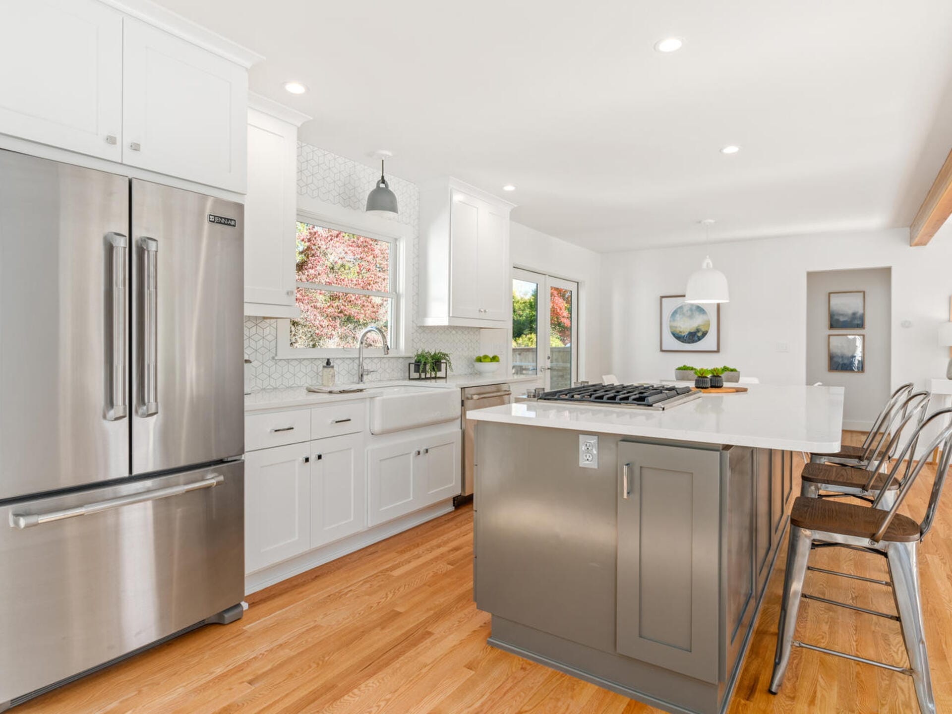 A modern kitchen with white cabinets, a stainless steel refrigerator, and a large island with a stovetop. Three metal bar stools are at the island. Hardwood floors and a large window bring in natural light.