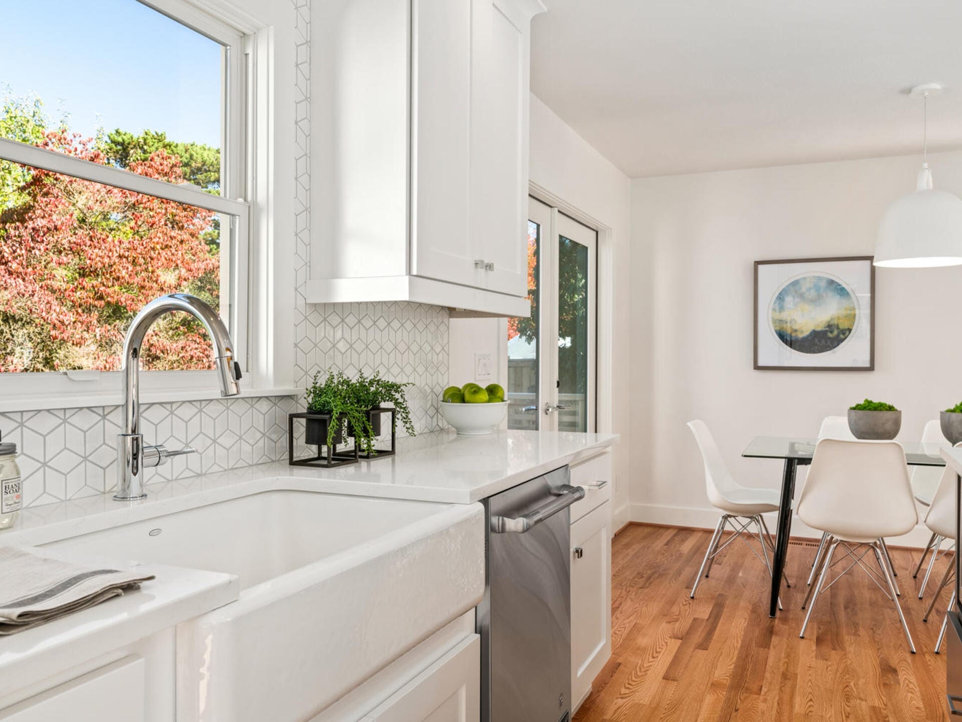 A bright kitchen with white cabinets and countertops, featuring a farmhouse sink and stainless steel dishwasher. A dining area with a table and white chairs is in the background. A window shows trees with autumn foliage outside.