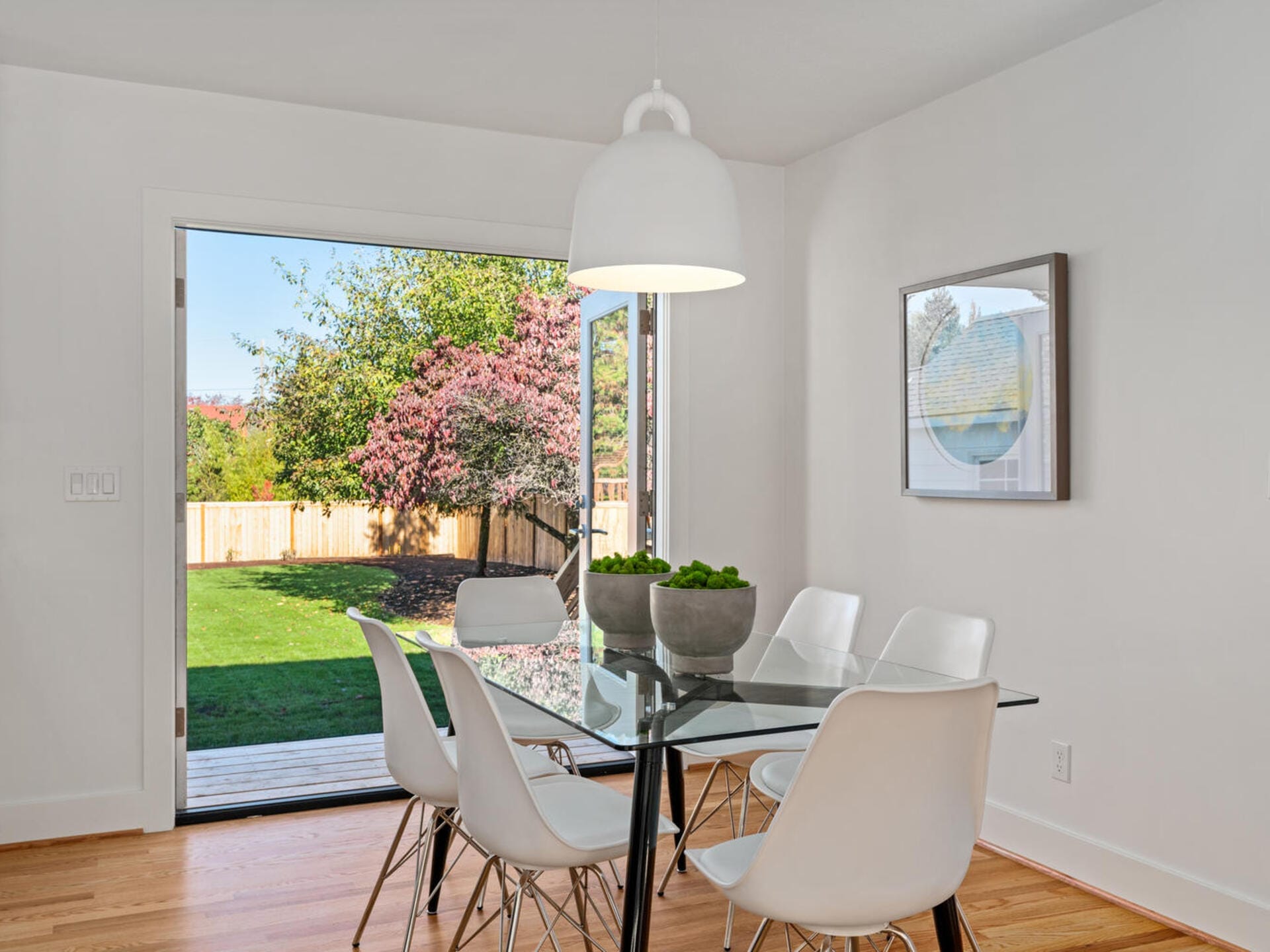 A modern dining room with a glass table and six white chairs. A bowl of green apples sits on the table. A large window opens to a garden with a wooden fence and colorful trees. A white pendant lamp hangs above the table.