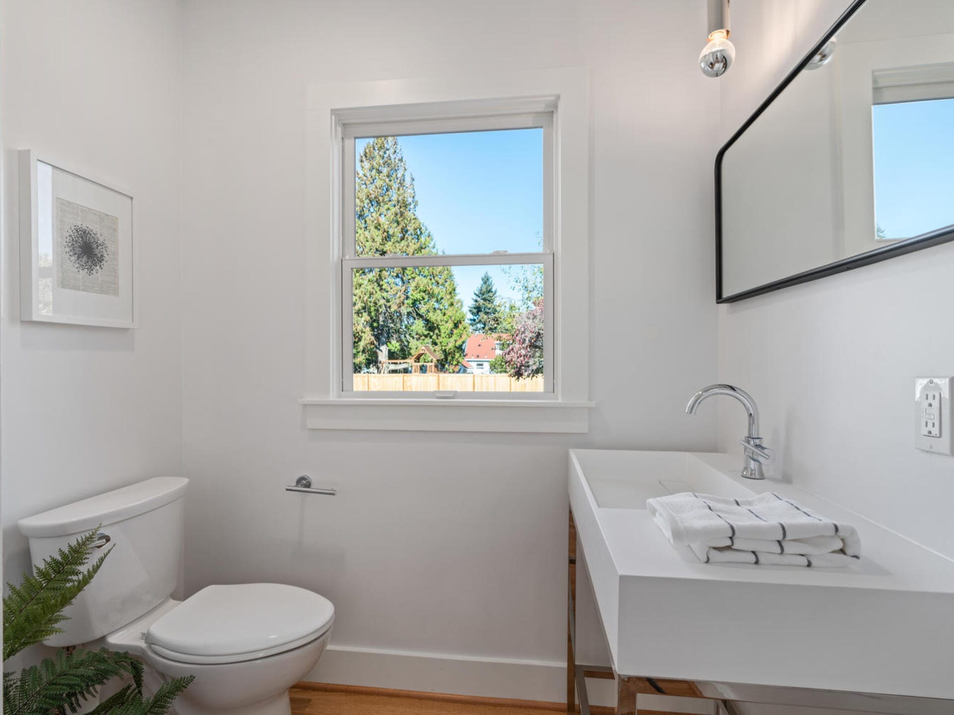 A bright bathroom featuring a white toilet, a sleek white vanity with a modern faucet, and a large rectangular mirror. A window provides natural light, and a framed picture and green plant add decor.