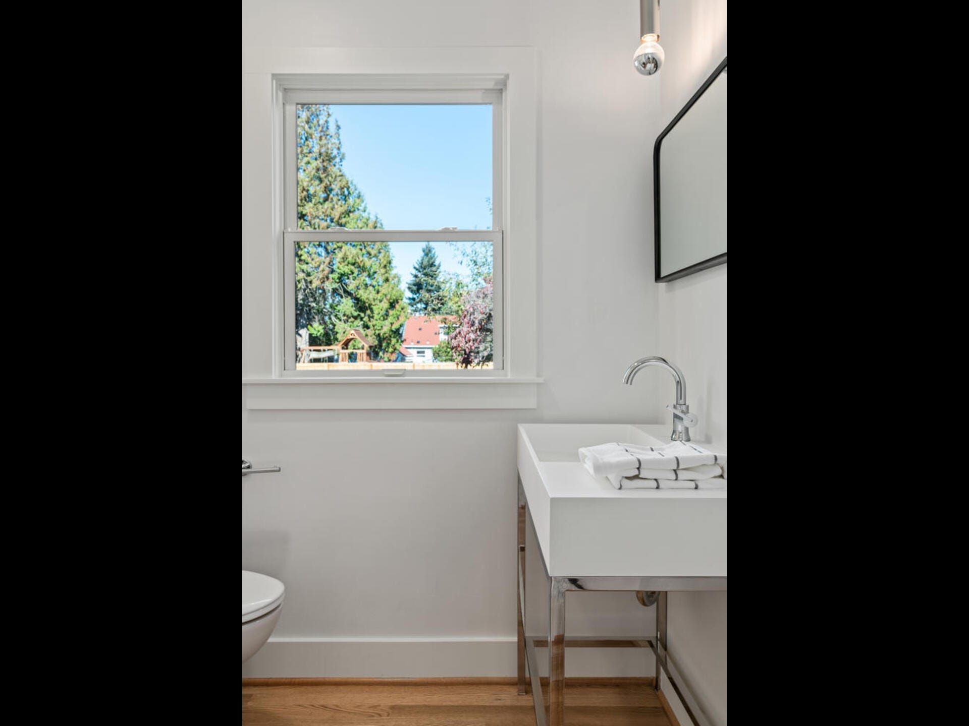 A minimalist bathroom with a white sink and chrome faucet, a small stack of white towels, a wall mirror, and a window showing a view of trees and a house. The room has white walls and a light wooden floor.