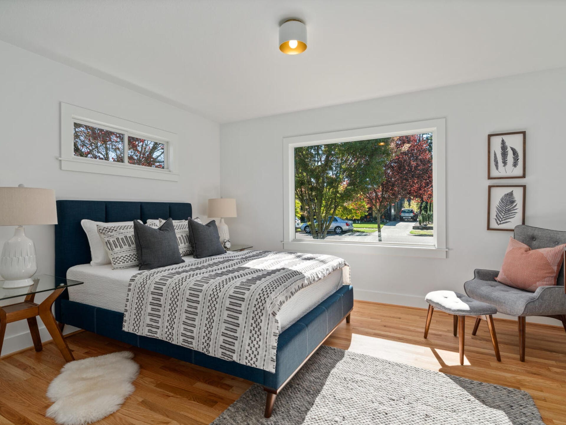 A bright bedroom with a large window showing a tree-lined street. The room features a bed with patterned bedding, a gray upholstered chair with a pink pillow, and two framed leaf artworks on the wall. A furry rug lies on the wooden floor.