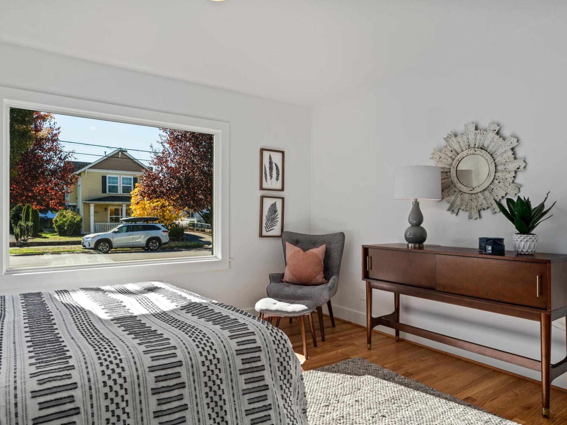 A bright bedroom with a large window showing a suburban street. Inside, a bed with a patterned blanket, a gray chair with a cushion, a wooden dresser with a lamp, a plant, and a decorative mirror are visible on hardwood floors.