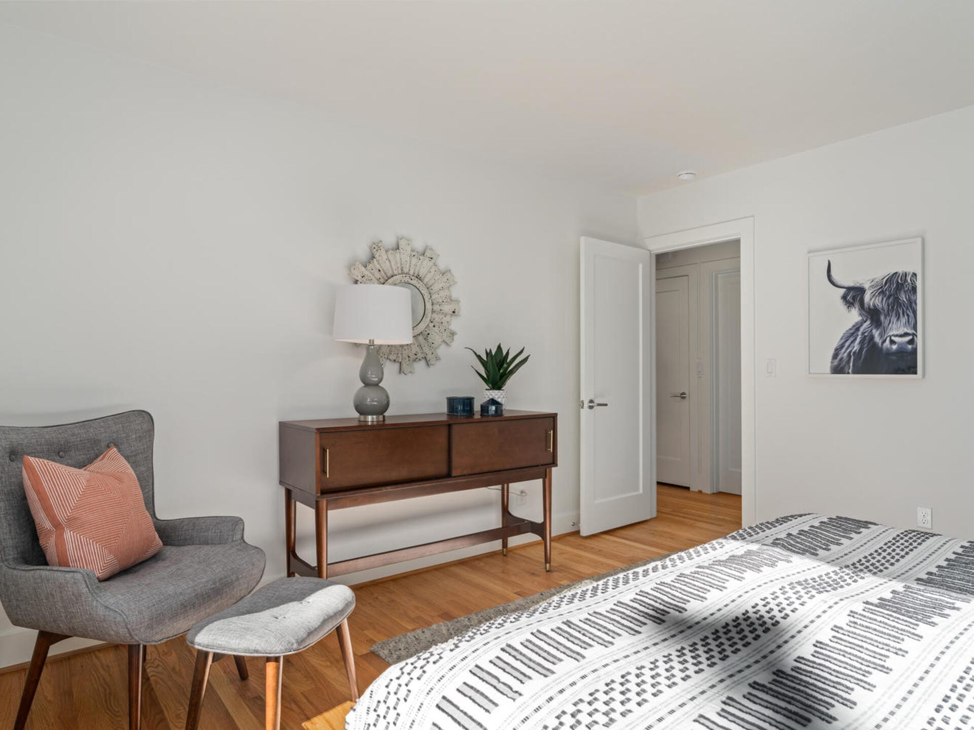 A bright, minimalist bedroom with a geometric-patterned bedspread. A mid-century chair and footstool sit next to a wooden dresser with a mirror, lamp, and plant. An open door reveals a hallway, and a framed bull artwork hangs on the wall.