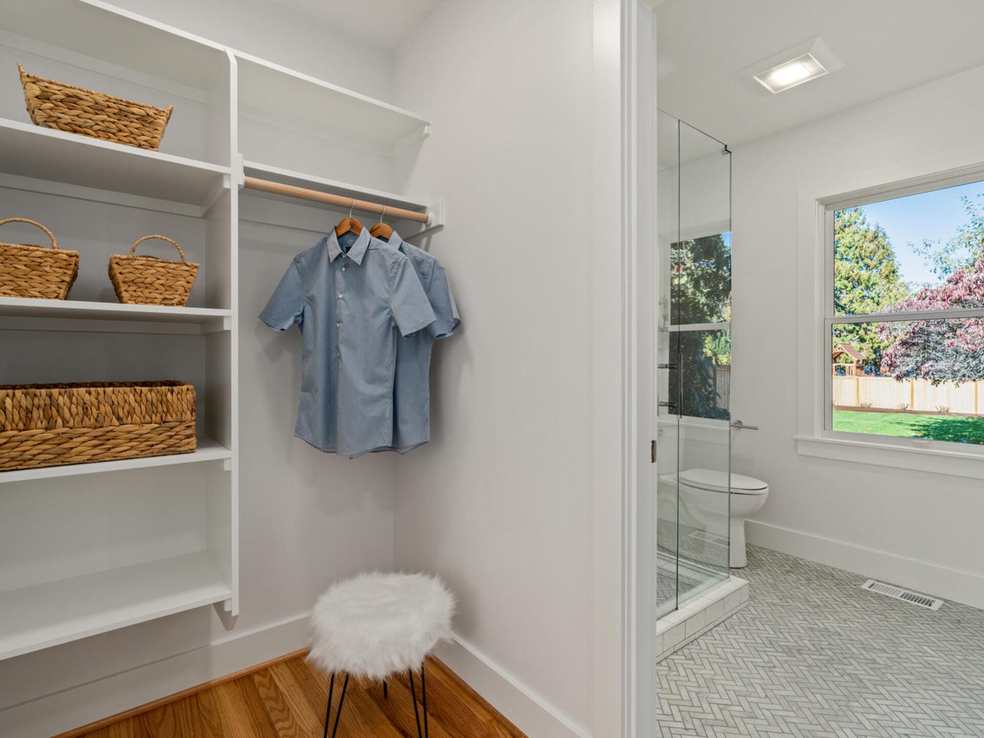 A neatly organized walk-in closet with white shelves, baskets, and a hanging blue shirt. A fluffy stool sits on a wooden floor. The bathroom with a glass shower and a window is visible in the background.