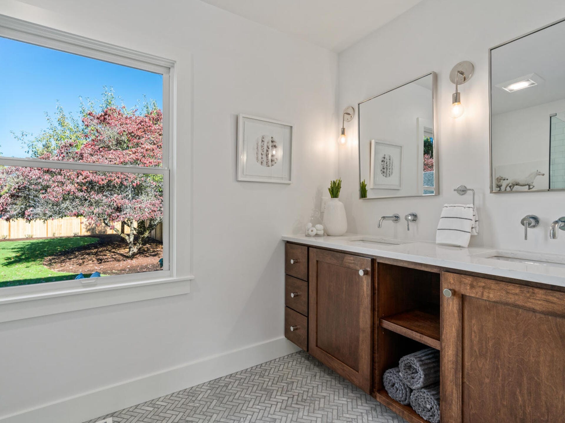 A modern bathroom with a large wooden vanity featuring dual sinks and mirrors. The countertop holds rolled towels and a small plant. A window reveals a view of a tree and a wooden fence outside. The floor has a chevron-patterned tile.