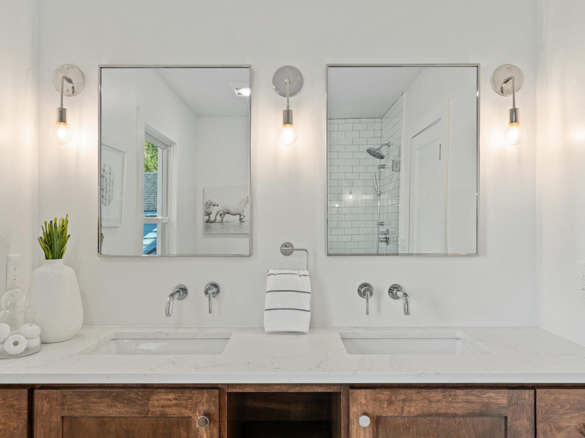 A modern bathroom features two square mirrors above double sinks with silver faucets. A stack of white towels is centered between the sinks. The walls are white, and two light fixtures with exposed bulbs are above the mirrors.