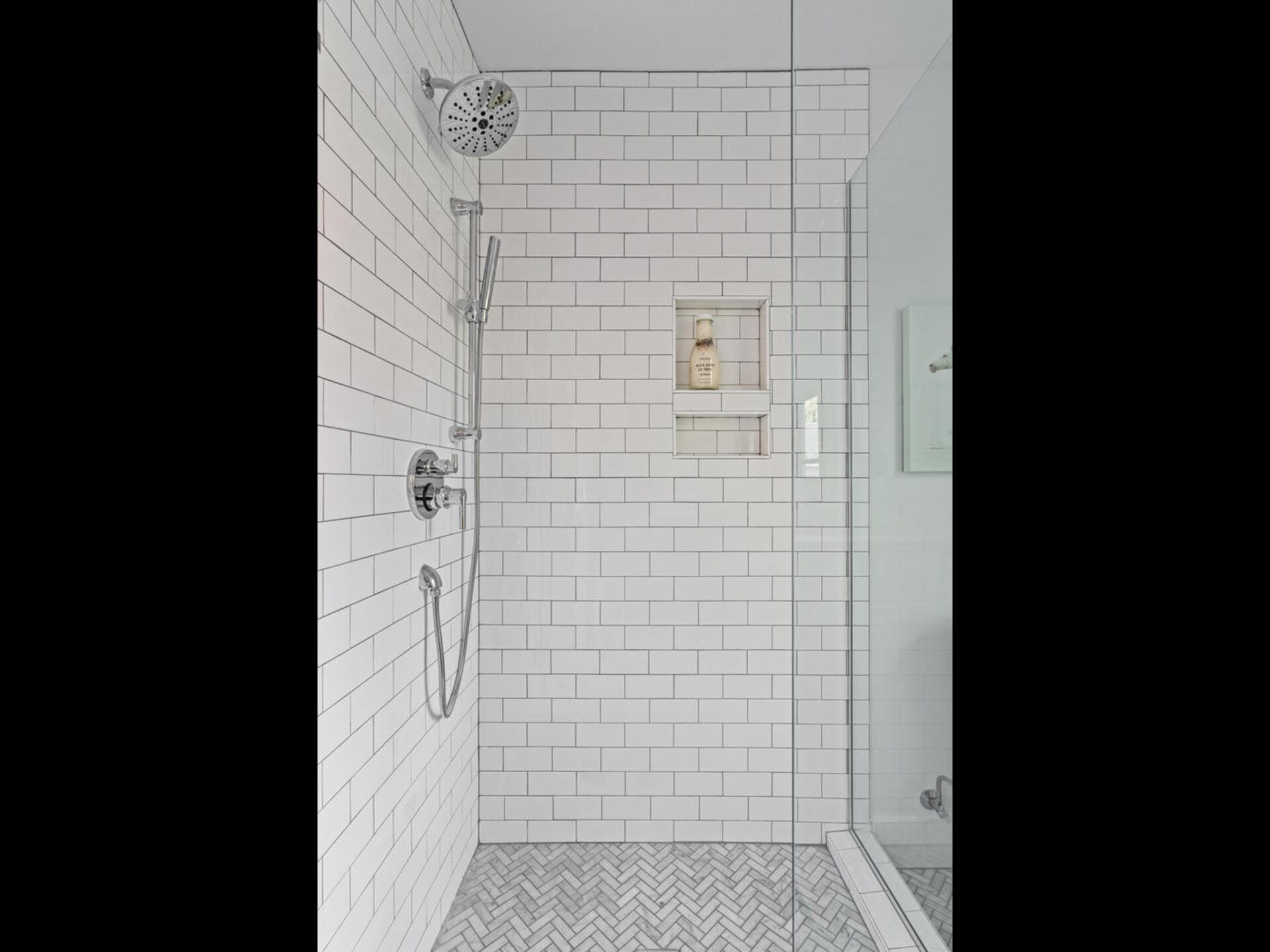 Modern shower with white subway tiles, a wall-mounted adjustable showerhead, and a handheld sprayer. A recessed shelf holds toiletries. The floor has a herringbone tile pattern, and a glass door encloses the space.