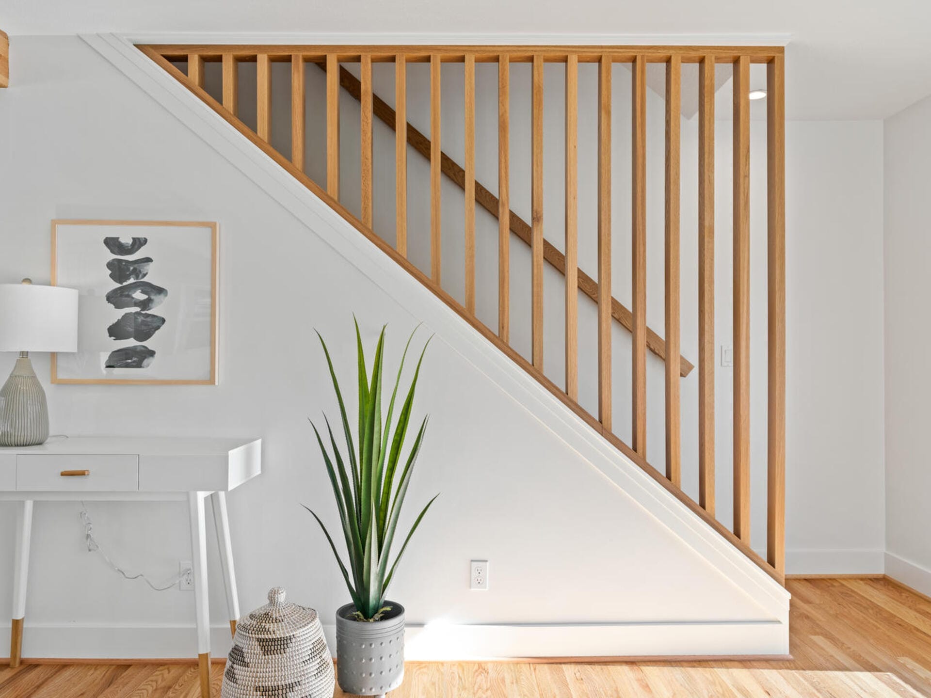 A modern interior featuring a slatted wooden staircase with white walls. A green plant in a pot sits on hardwood flooring alongside a woven basket and a white table with a lamp and framed artwork. The space is bright and minimalist.
