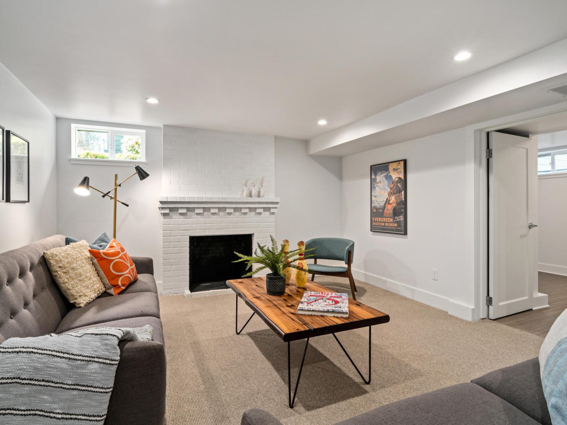 A cozy living room with a white brick fireplace, gray sofas, a wooden coffee table with a potted plant, and a framed poster on the wall. The room is well-lit with modern ceiling lights and has small windows near the ceiling.
