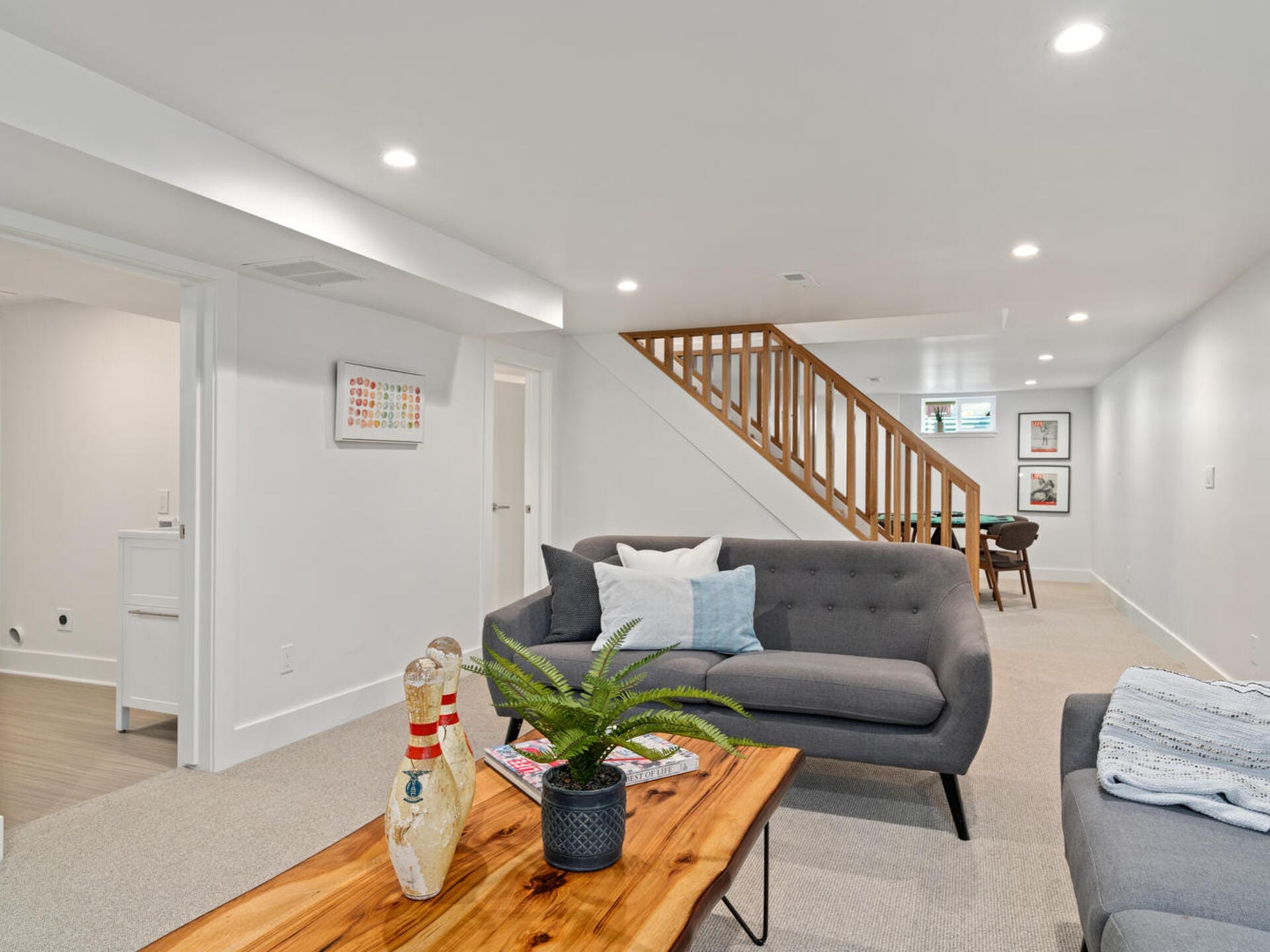 A modern living room with neutral tones features a gray sofa, a wooden coffee table with a fern and decorative items, and a staircase leading upward. Wall art decorates the space, and a laundry area is visible through an open door on the left.