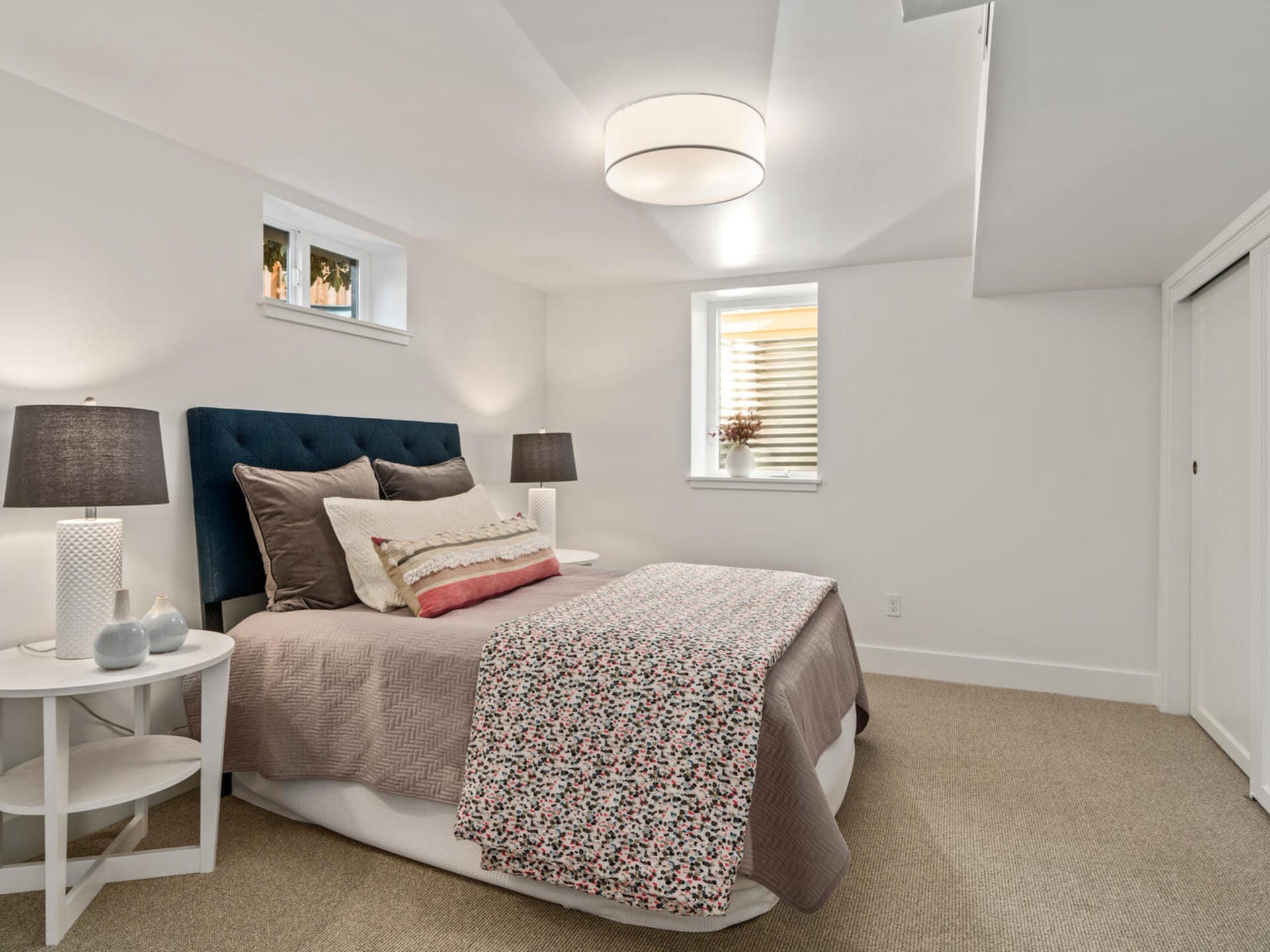A cozy bedroom with a double bed featuring a floral-patterned quilt and multiple pillows. Two bedside tables with dark lamps flank the bed. A round ceiling light illuminates the room, which has a window with closed blinds and light-colored walls.
