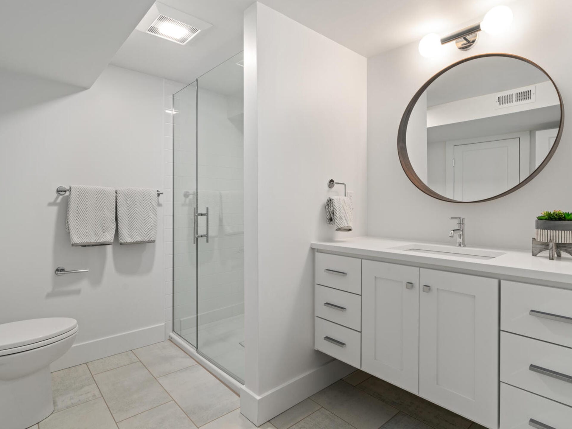 Modern bathroom with a glass-enclosed shower, white vanity with a round mirror, and a white countertop. Towels hang on the wall, and a potted plant decorates the countertop. The space is bright with overhead lighting.