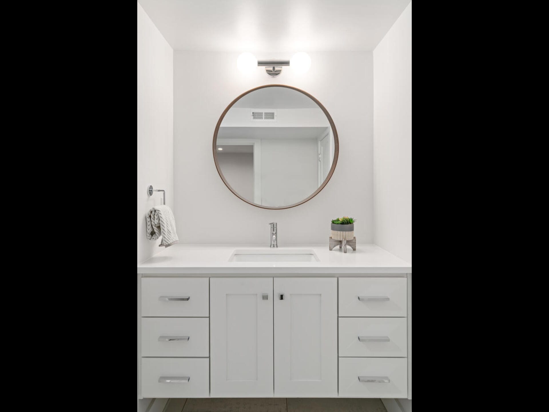 A minimalist white bathroom features a round mirror above a sink with a white countertop. The vanity has several drawers, and a small potted plant adds a touch of greenery. A towel is neatly draped on a rack to the left.