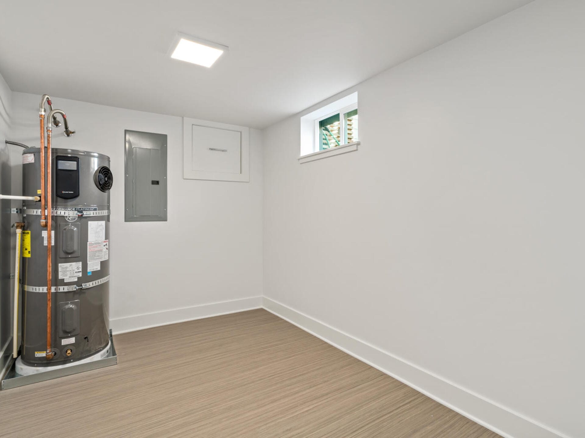 A clean, empty utility room with light wood flooring. It contains a water heater on the left, and a small window near the ceiling on the right. The walls are plain and white, giving a minimalist appearance.